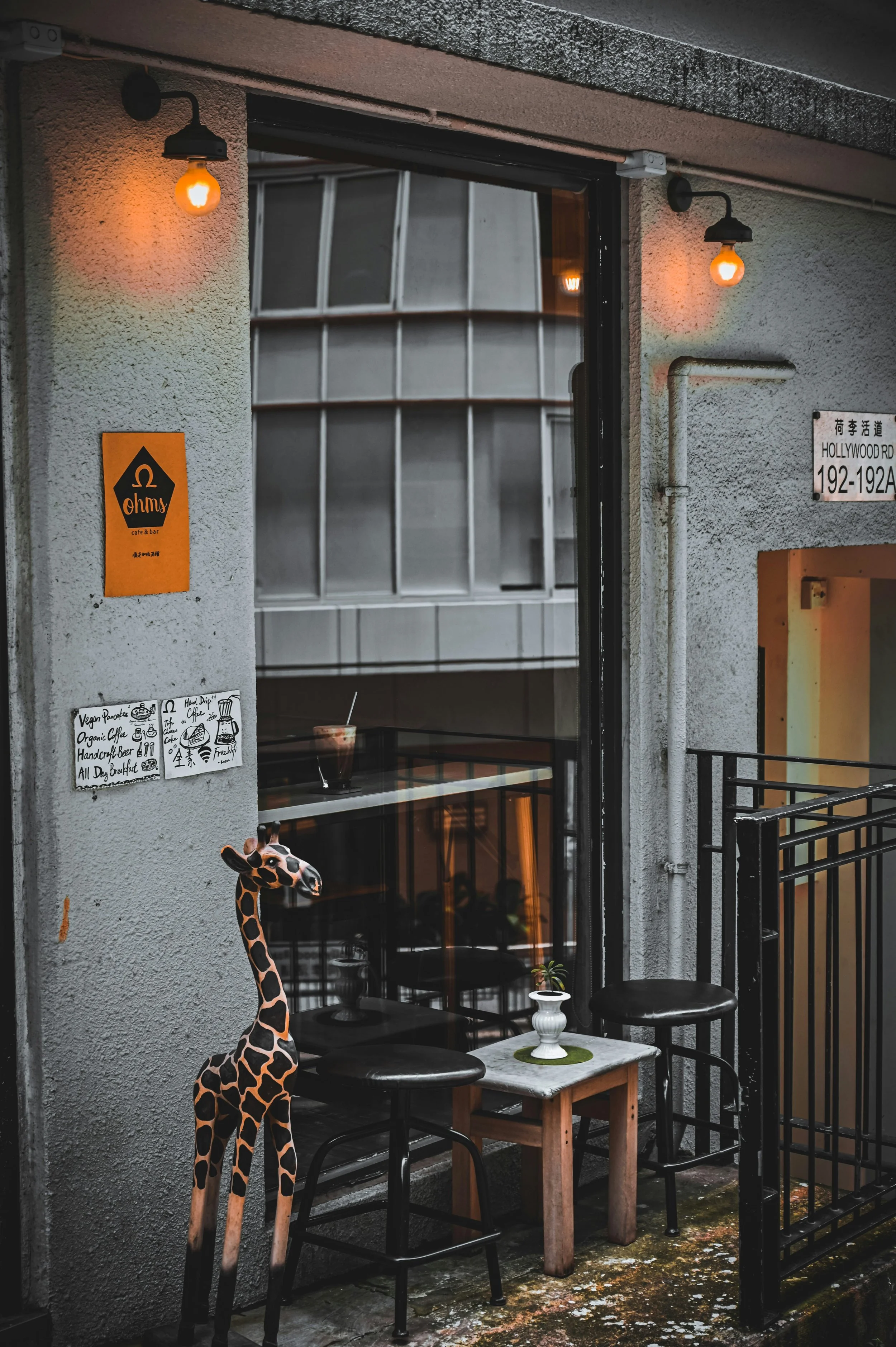 A small outdoor seating area next to a cafe or bar with two black chairs, a small wooden table, and a small potted plant with a giraffe-shaped plush toy leaning against a chair. The facade has a few wall-mounted orange lights, and signs indicating the location on Hollywood Road and Ohms cafe & bar. The ground appears slightly dirty and mossy.