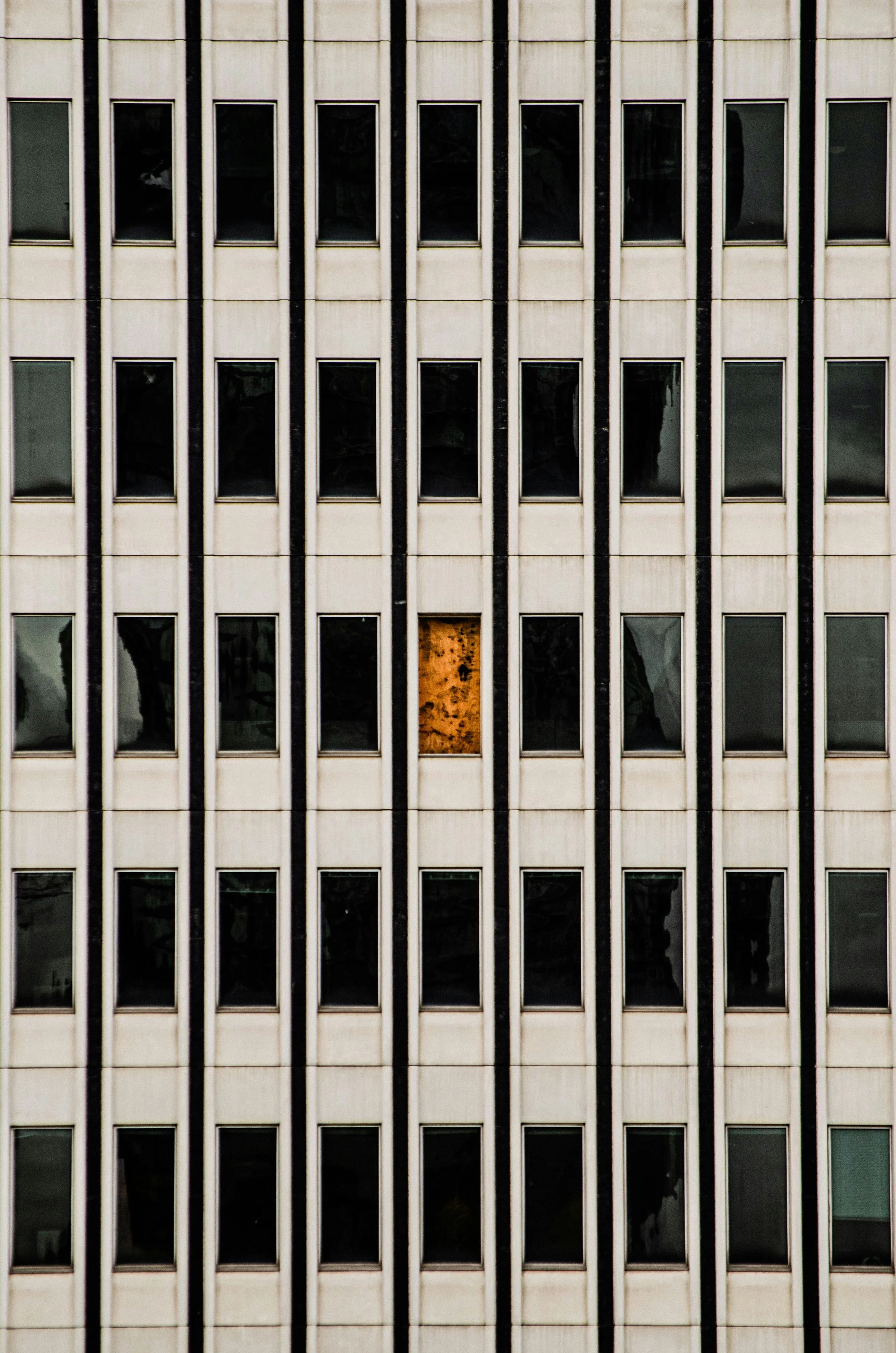 The image shows the exterior of a high-rise office building with a grid of rectangular windows, some of which are dark and some are reflecting trees or the sky. One window in the bottom row is boarded up with plywood, showing signs of rust around the edges.