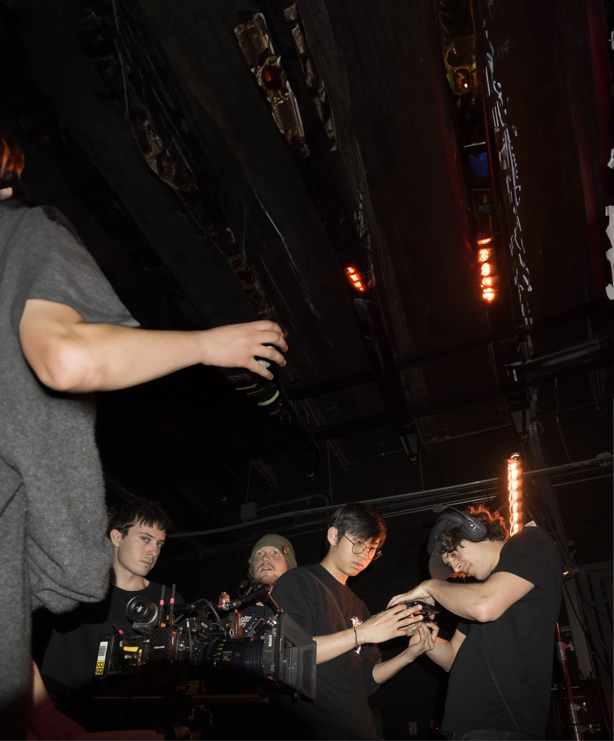 A group of people working behind the scenes at a film or video production, with camera equipment and a monitor displaying footage.
