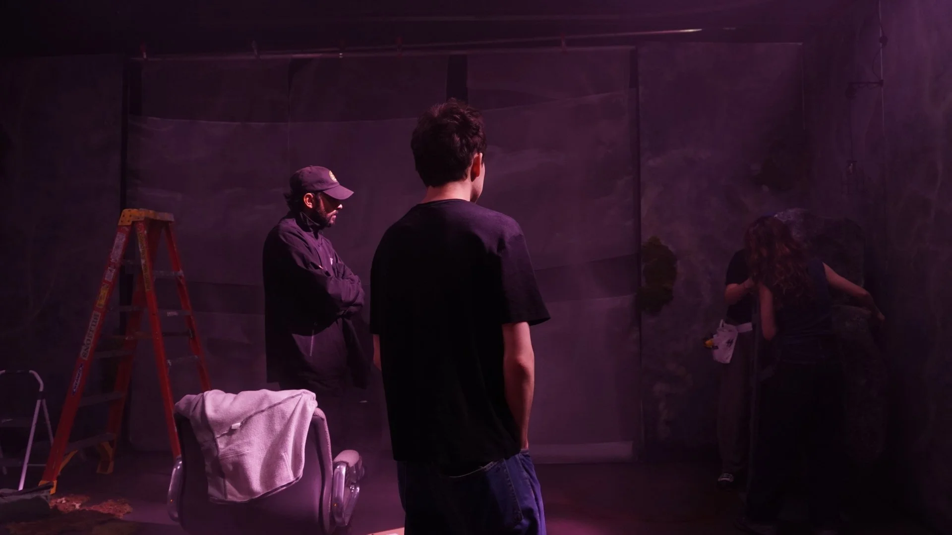 People working on a set with a ladder, a chair with a towel, and a person adjusting a large backdrop in dim purple lighting.