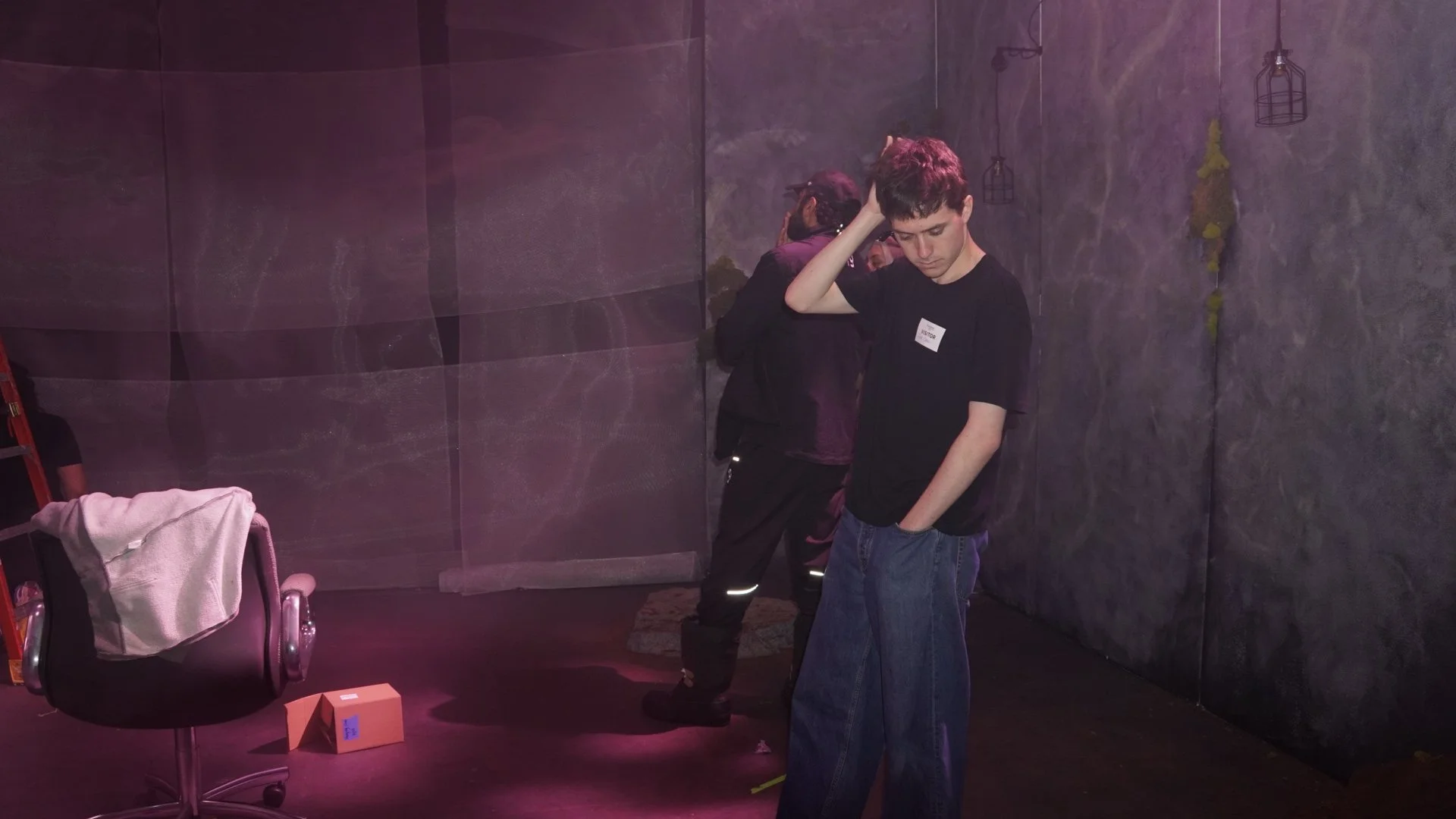 Young man in black T-shirt and blue jeans standing, looking down, hand on head, in dark room with gray textured walls, two hanging light fixtures, a firefighter in uniform in background, black office chair with a gray cloth draped over it, orange box on floor.