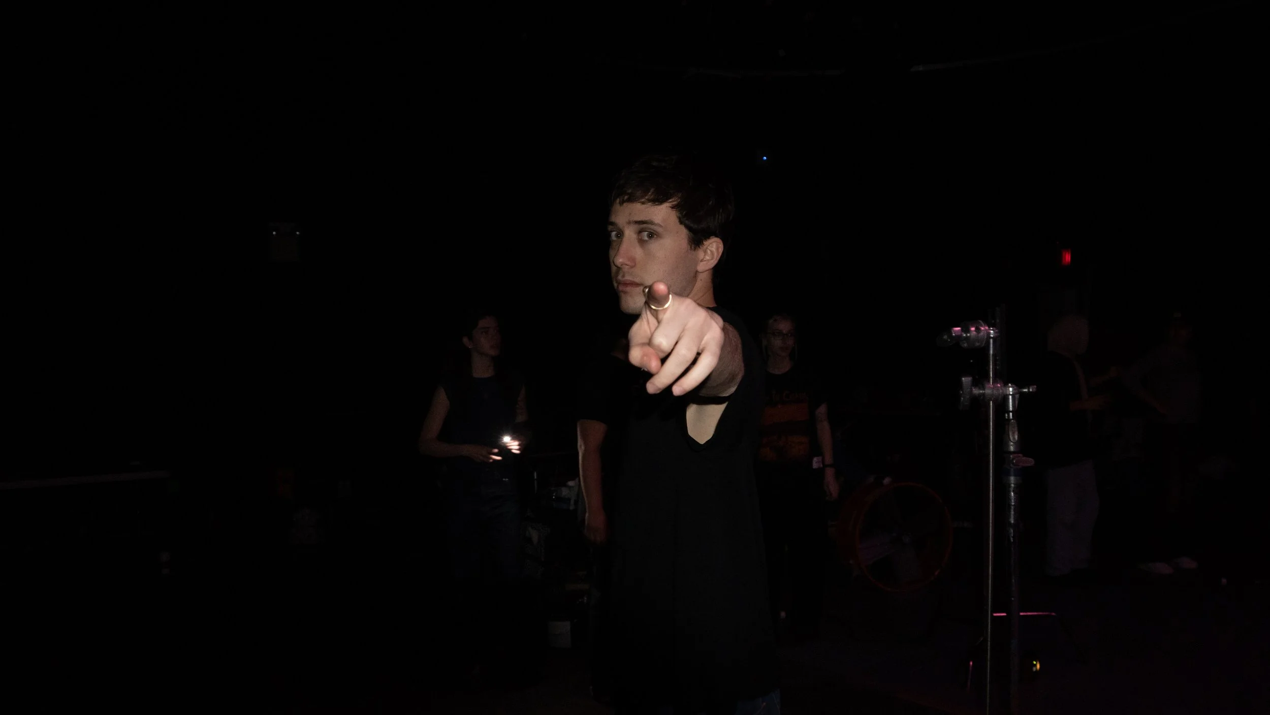 A young man in black clothing pointing towards the camera, with other people faintly visible in a dark setting.