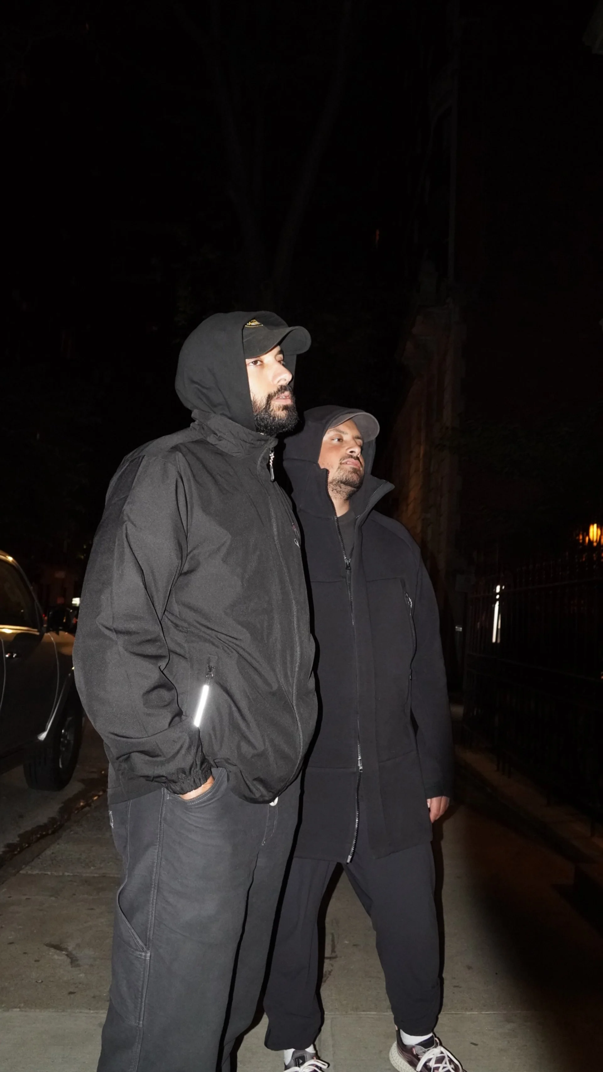Two men stand on a sidewalk at night wearing dark hooded jackets and caps, with serious expressions, next to parked cars and a dark building.