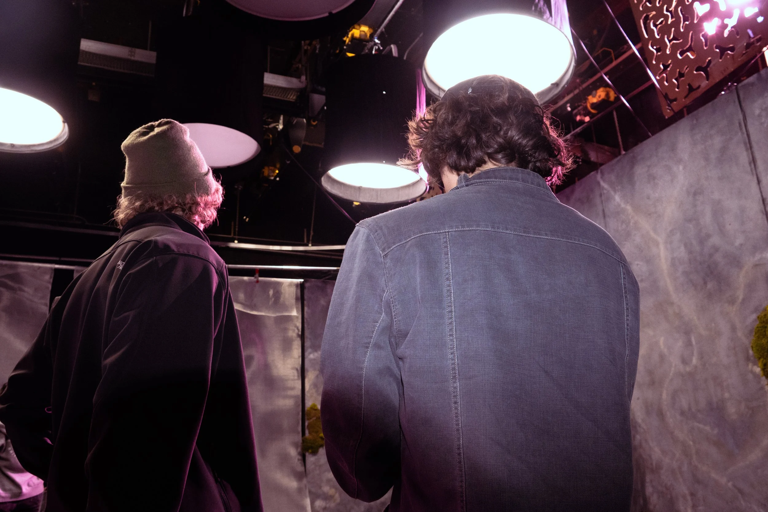 Two young men standing in a room with purple lighting, one wearing a beanie and black jacket, the other with curly hair and a denim jacket, facing away from the camera.