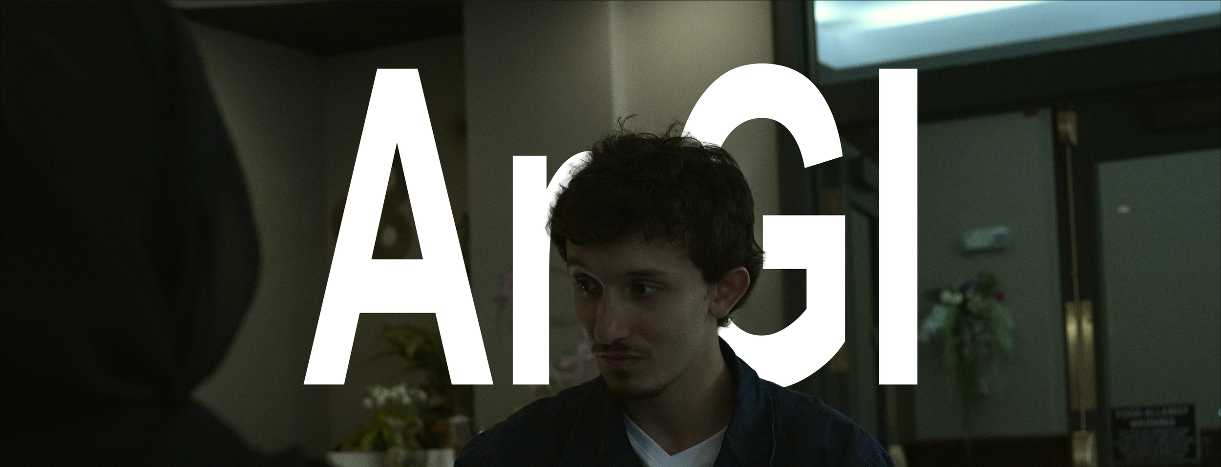 A young man with dark hair and a light beard looking to the side inside a building with a glass door in the background. Large white text reading 'A G' overlays the image.