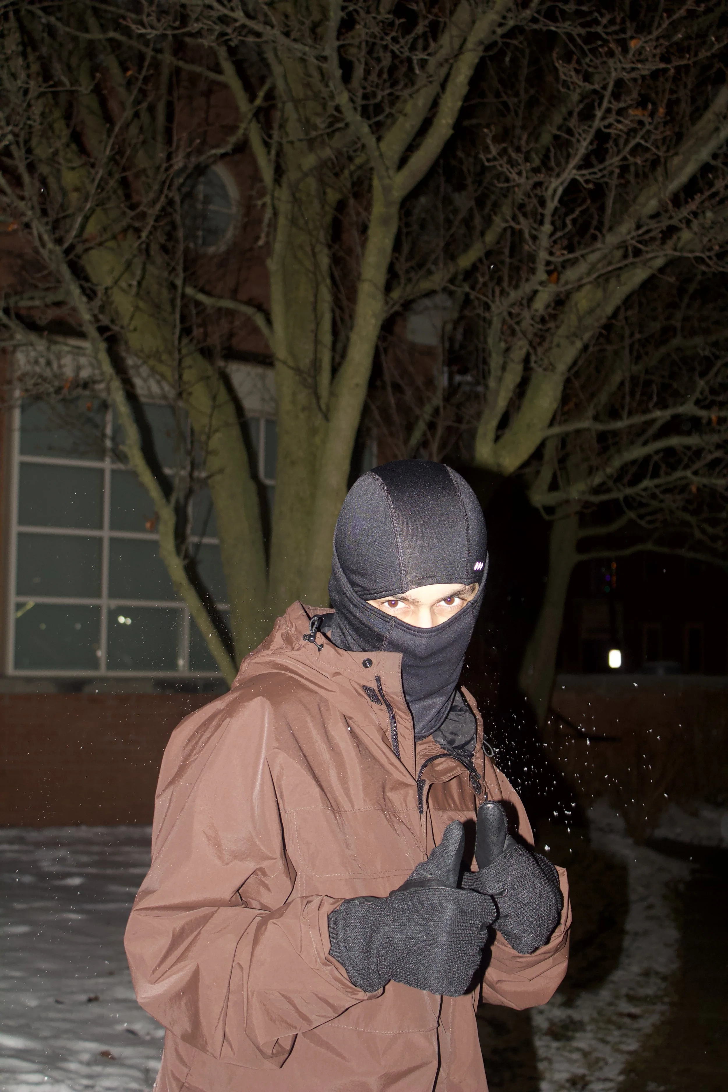 Person wearing a black balaclava, brown jacket, and gloves, giving two thumbs up outdoors at night.
