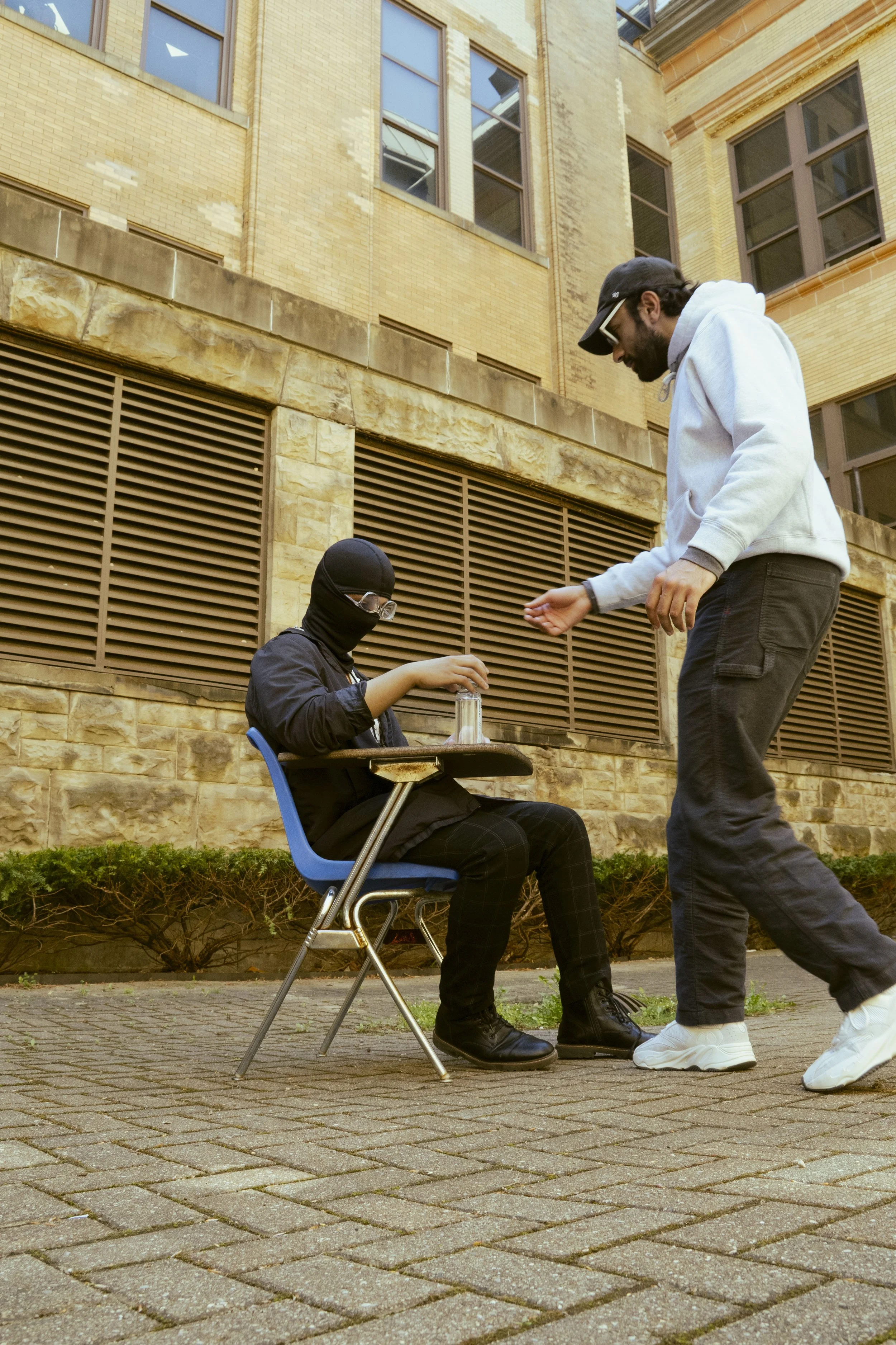 A man wearing a hoodie and sunglasses is handing money to a woman in a ski mask and glasses, who is sitting on a chair with a small table, outside near a brick building.