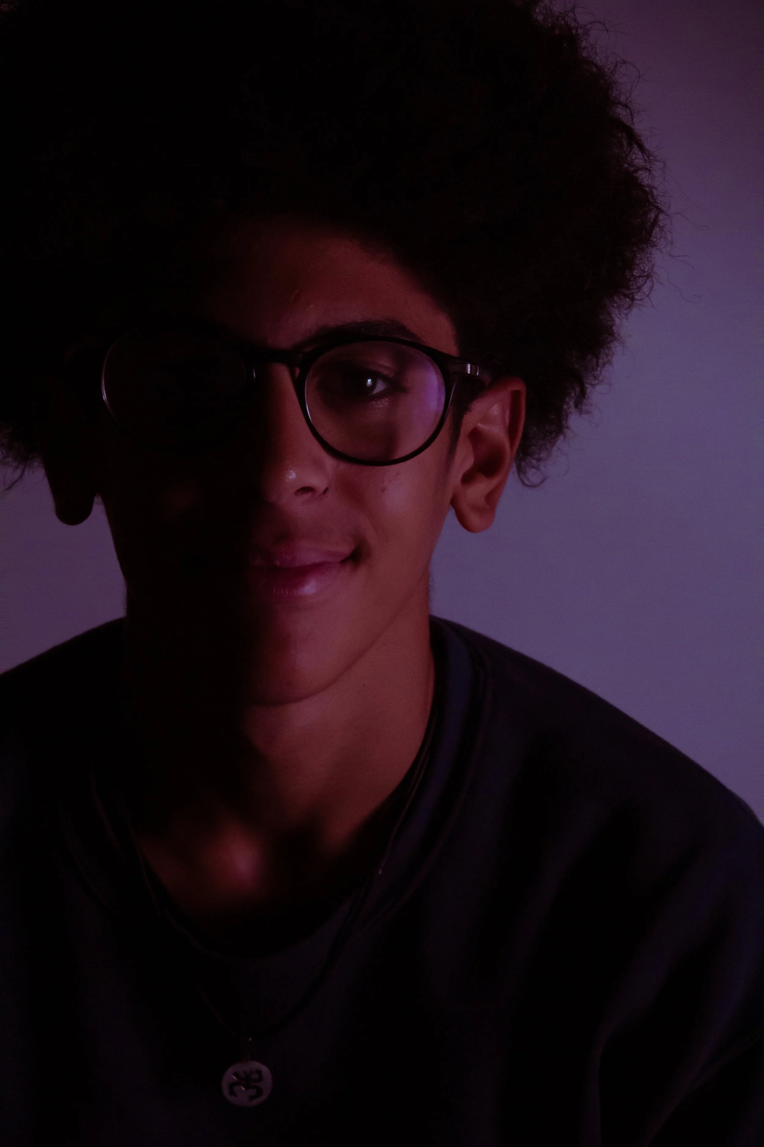 A person with glasses and an afro hairstyle smiling in dim, purple-toned lighting.