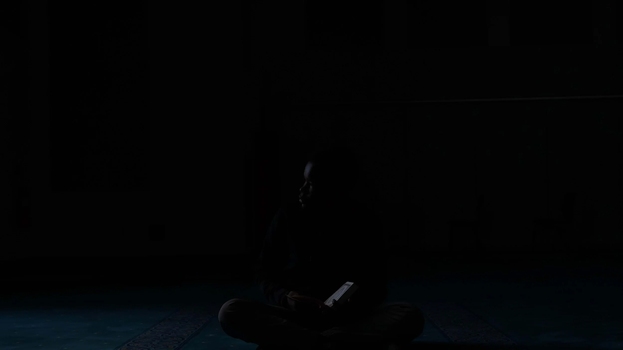 A person sitting cross-legged on a prayer mat in a dark room, illuminated by the light from a small electronic device they are holding.