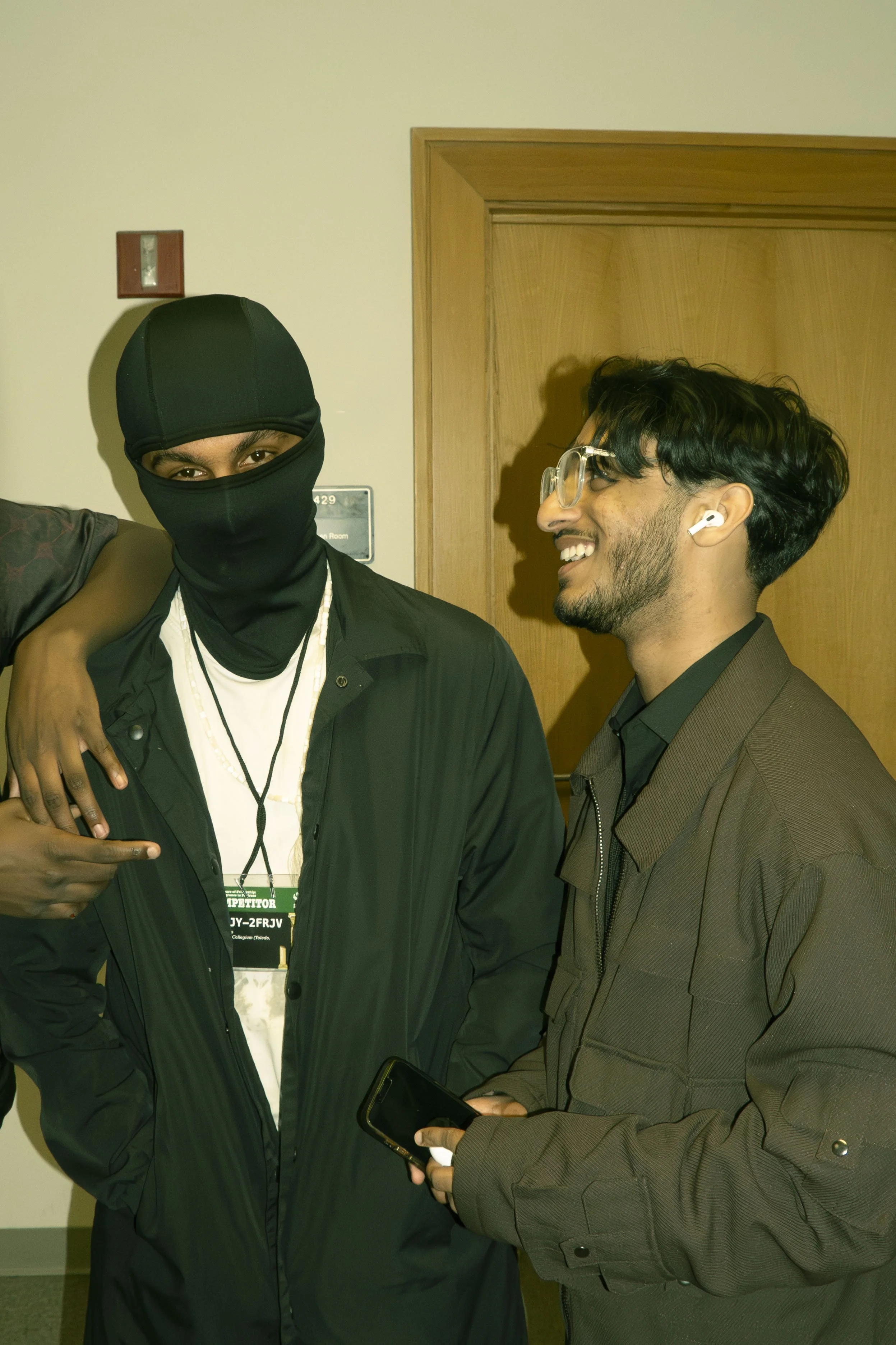 Two young men are smiling and conversing indoors, one wearing a black balaclava, black jacket, and white shirt with a lanyard, the other with glasses, earbud, and a brown jacket, holding a phone.