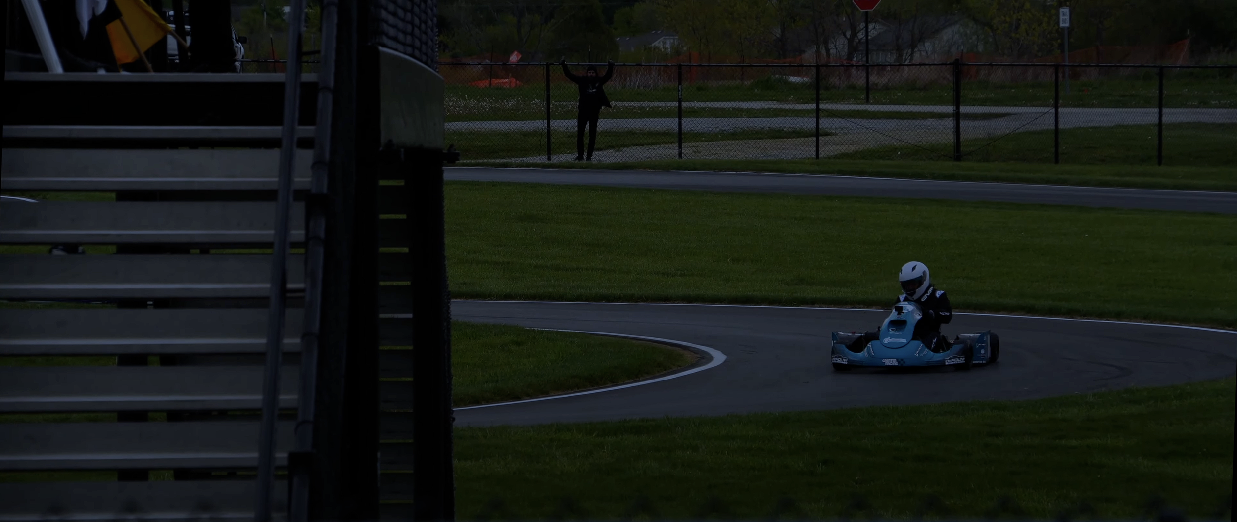 A person in a racing helmet driving a blue go-kart around a curve on a race track, with a spectator standing behind a fence with their arms raised in the background.