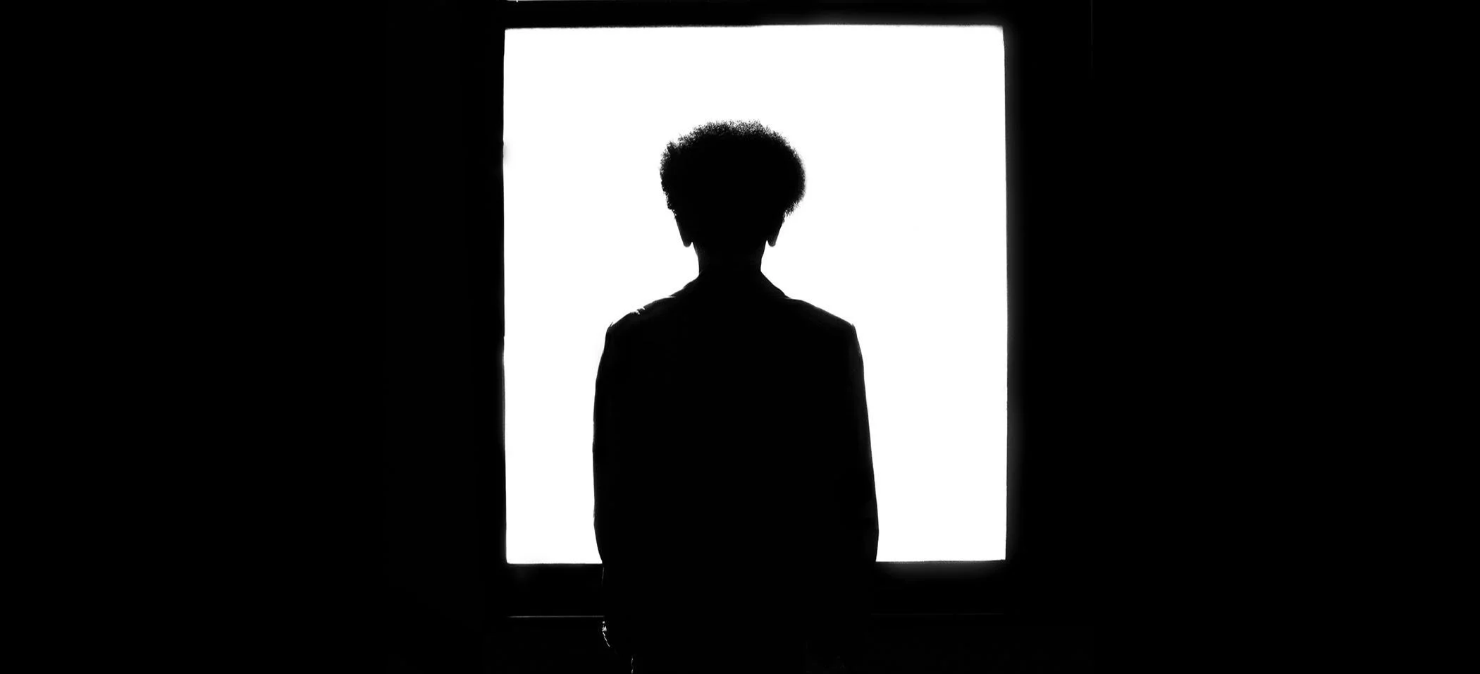 Silhouette of a person with curly hair standing and looking out a bright window.