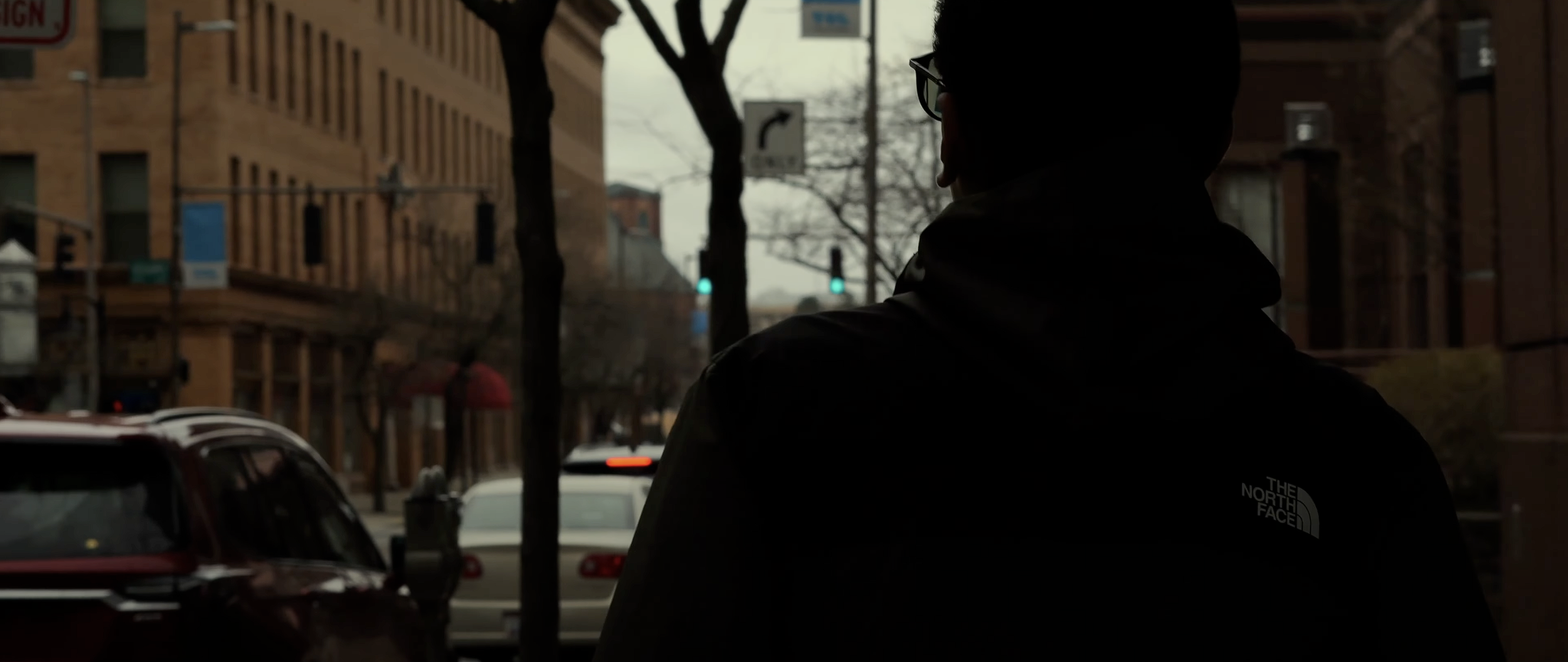 A silhouette of a person wearing glasses and a jacket with The North Face logo, standing on a city street during dusk or dawn, with cars and buildings in the background.
