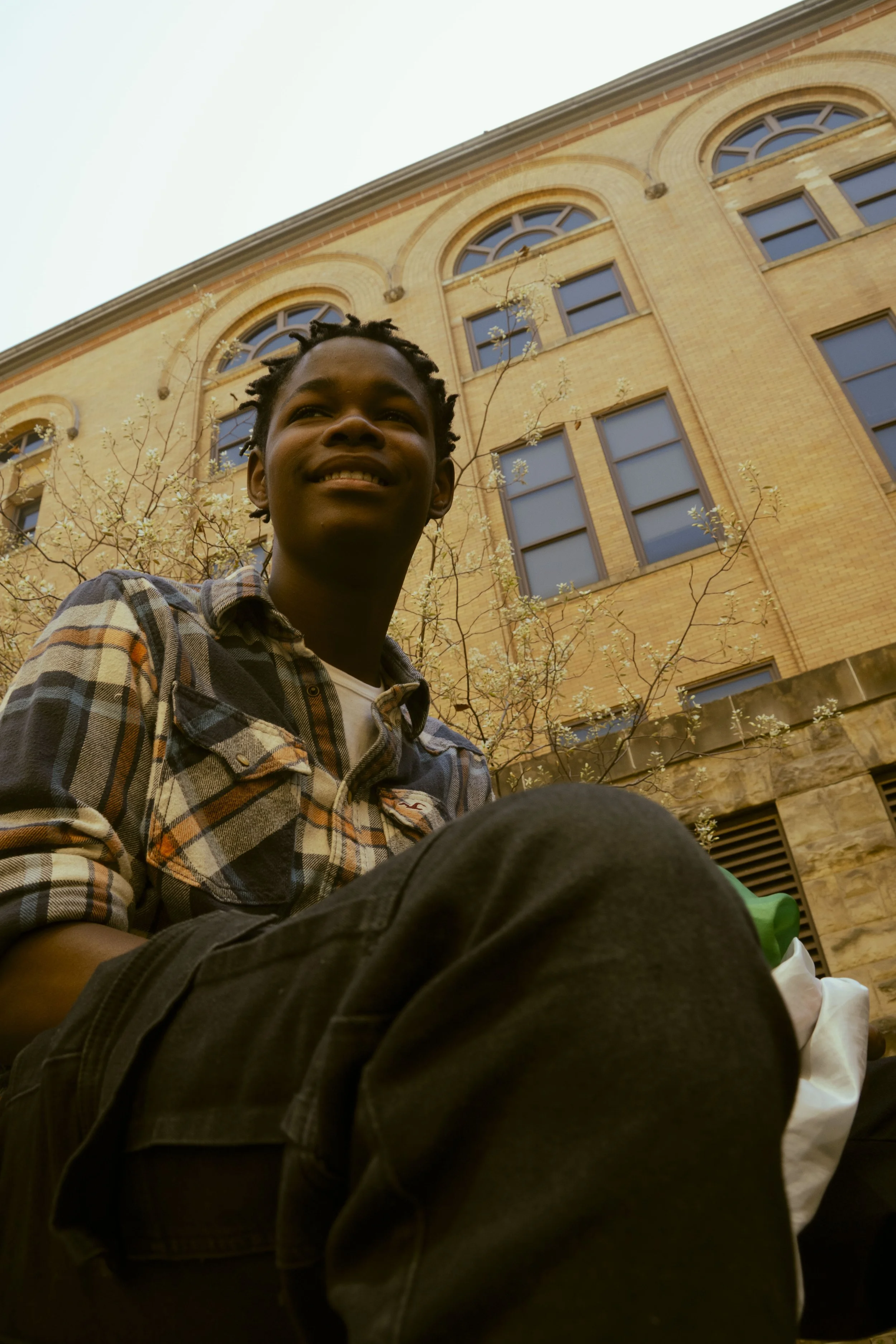 A young man with short dreadlocks, wearing a plaid shirt and black pants, sitting outside in front of a brick building with large arched windows and a tree with white blossoms.