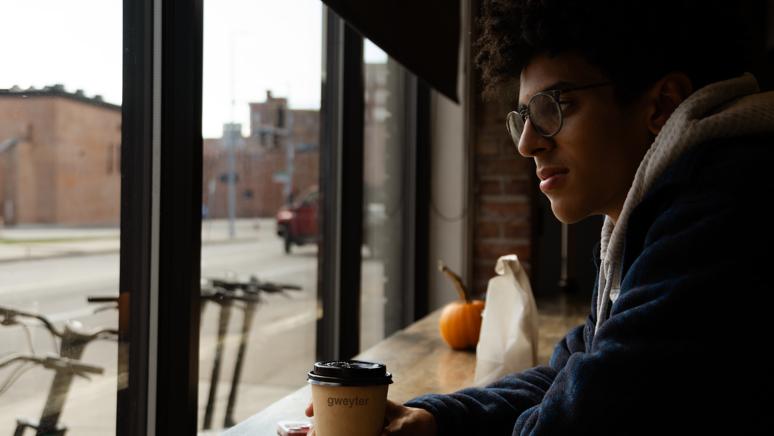 A young man with curly hair and glasses sitting at a wooden counter in a cafe, looking out the window. On the counter, there is a small pumpkin and a white paper bag. The man holds a to-go coffee cup labeled "Gweyter." Outside the window, a street sc
