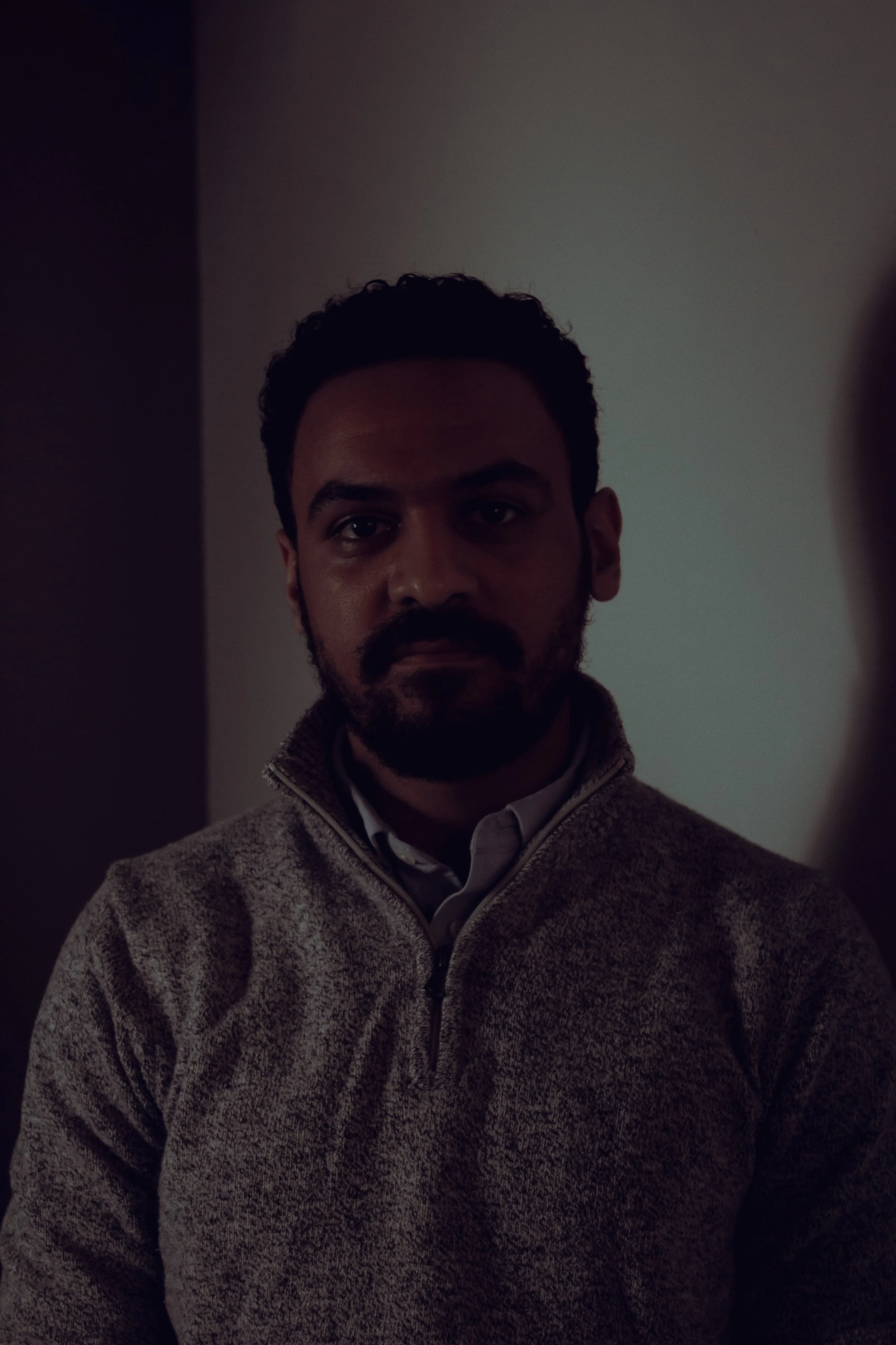 A man with dark curly hair and a beard looking at the camera, wearing a beige sweater.