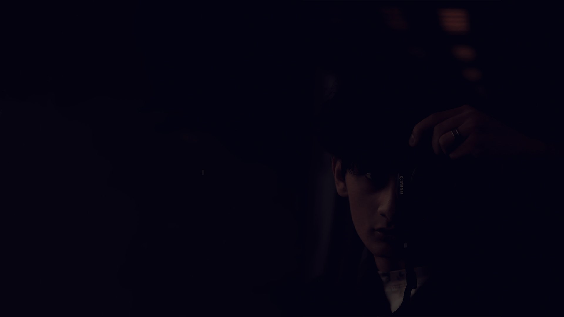 A person holding a camera in front of their face, surrounded by darkness, with only part of their face and hand visible.