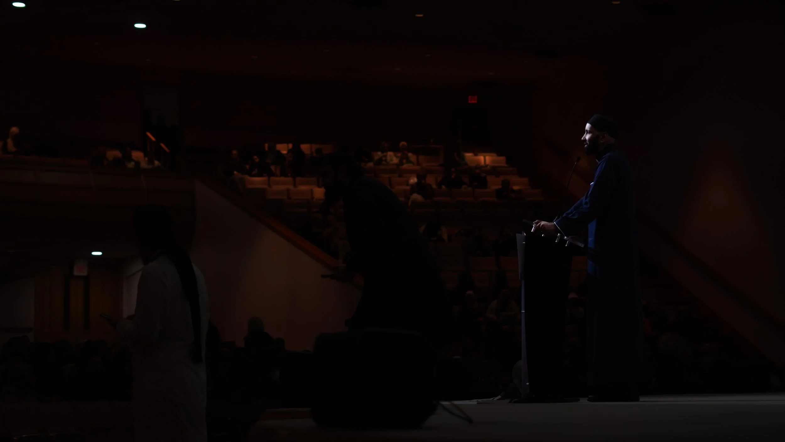 A man in a blue robe speaking at a podium on stage during a dark presentation. Audience members are sitting in the background, some lit slightly, and a person in a white coat uses a mobile phone in the foreground.