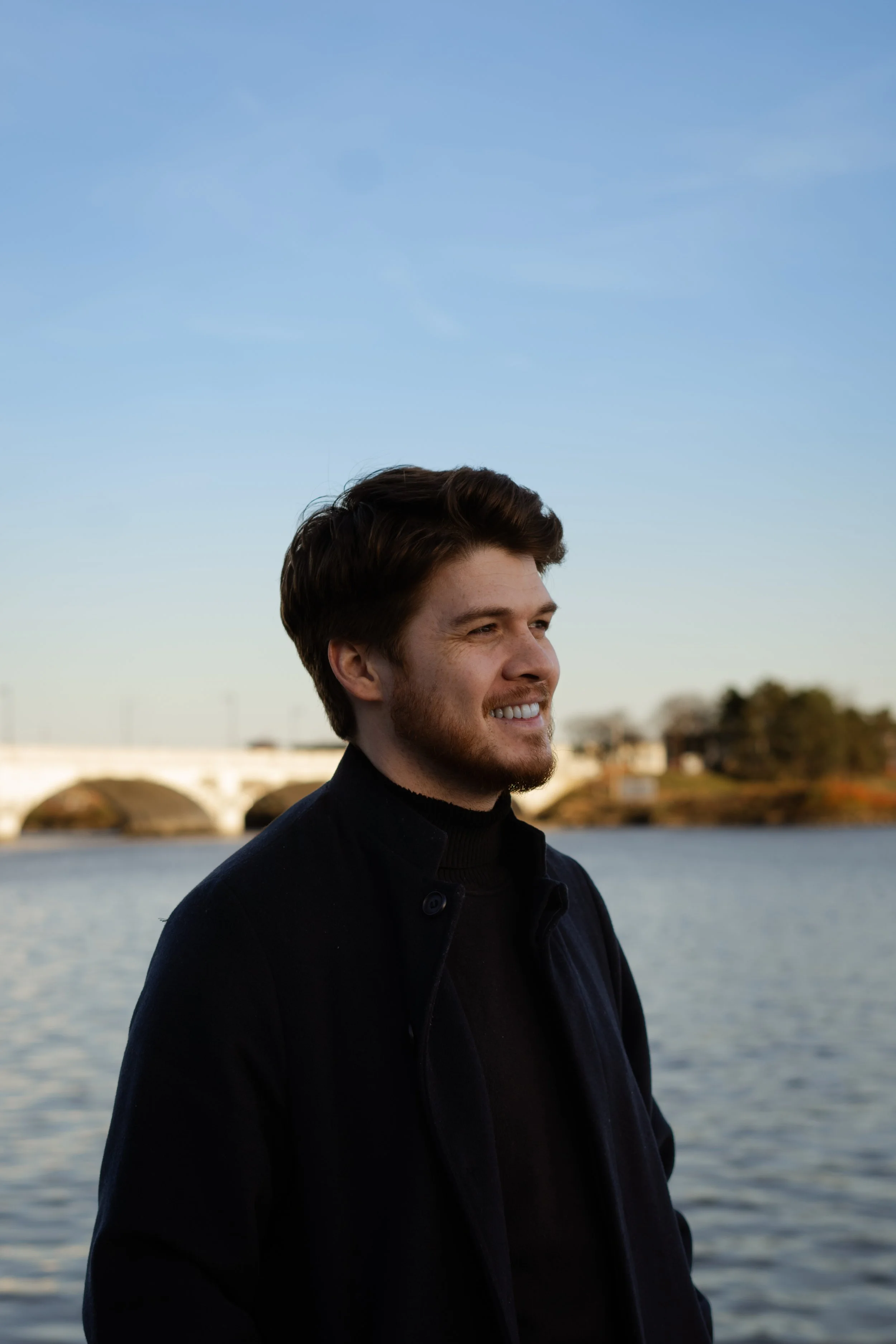 A young man with brown hair and a beard standing outdoors near a body of water, smiling, with a bridge and trees in the background under a clear blue sky.