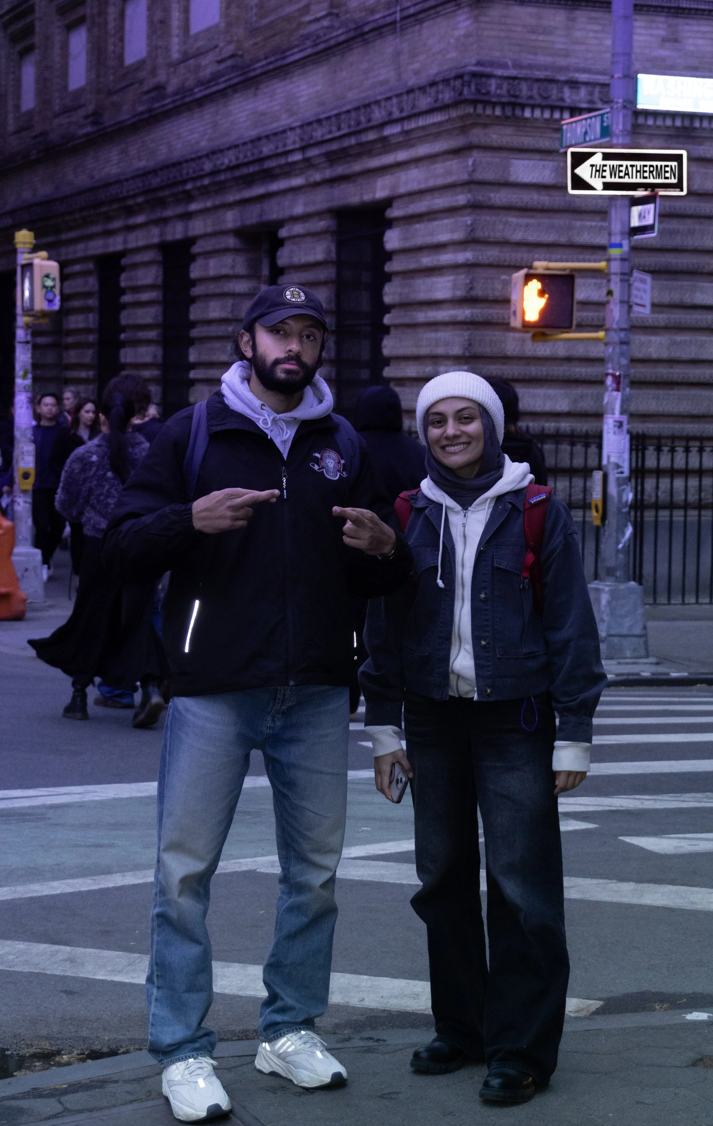 Two young people standing on a city street at dusk, with a large brick building and street signs in the background. The person on the left is pointing at the person on the right, both smiling.