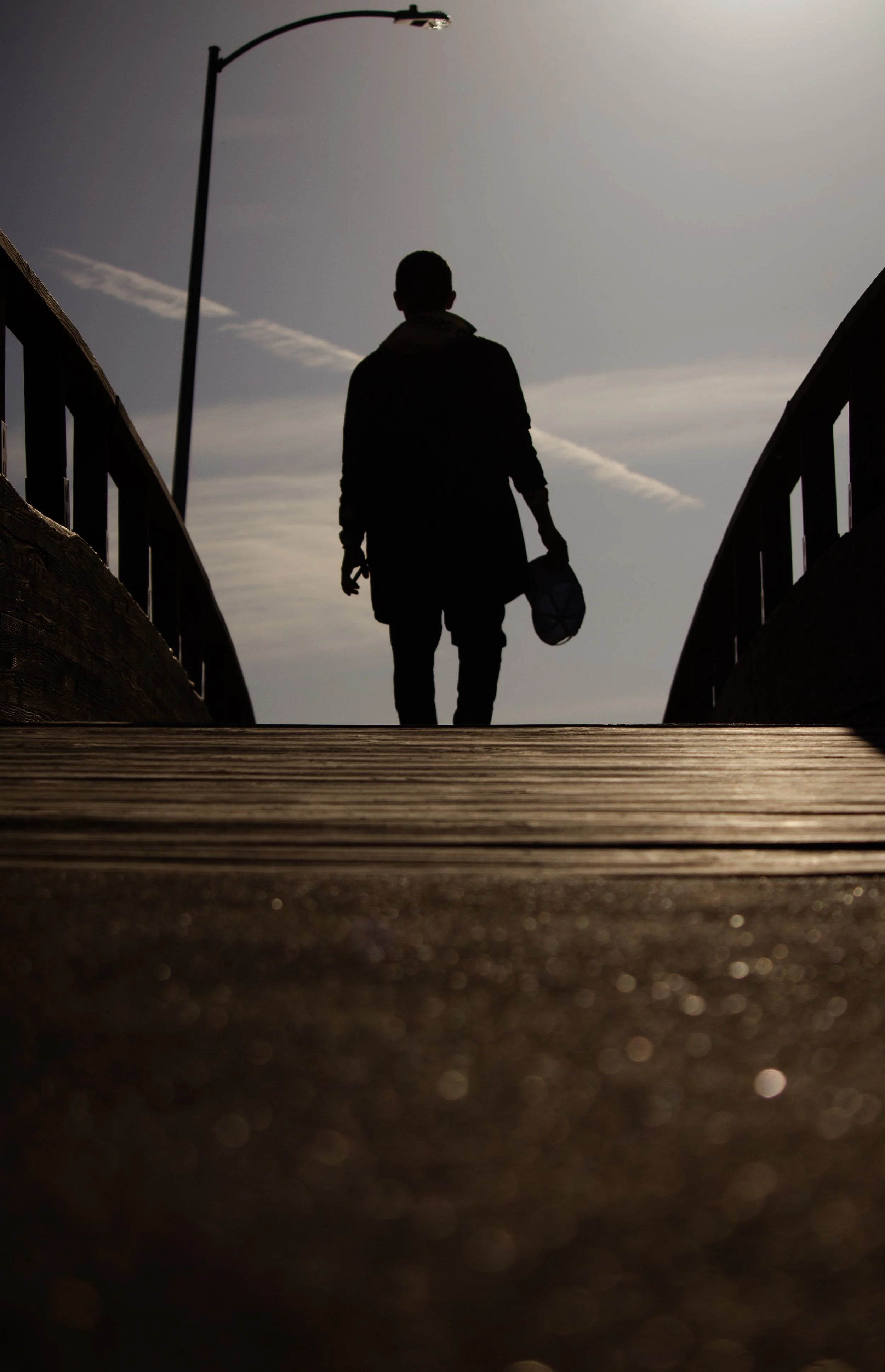 Silhouette of a man walking on a wooden bridge, holding a hat in his hand, with an overcast sky and a streetlight overhead.