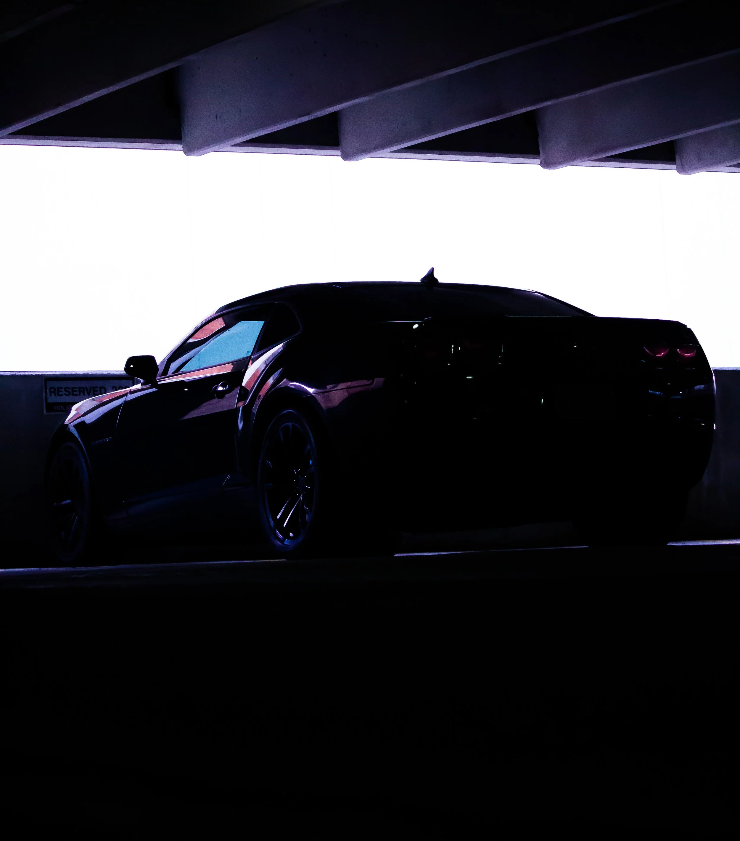 Dark-colored sports car parked in a parking structure with bright light behind it, silhouetting the vehicle.