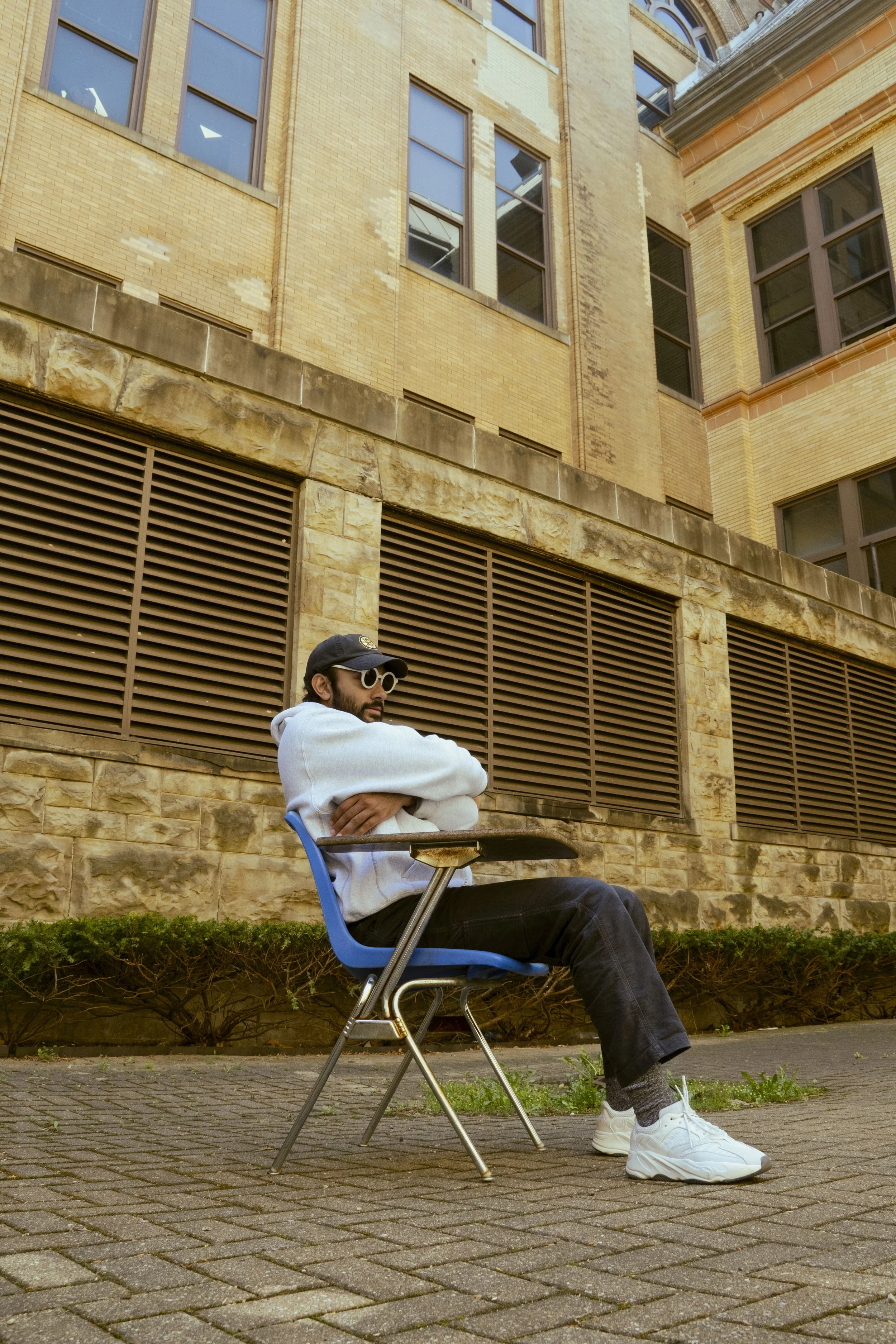 A man sitting on a blue plastic chair outdoors in front of a brick building, wearing sunglasses, a black cap, a white hoodie, black pants, and white sneakers.