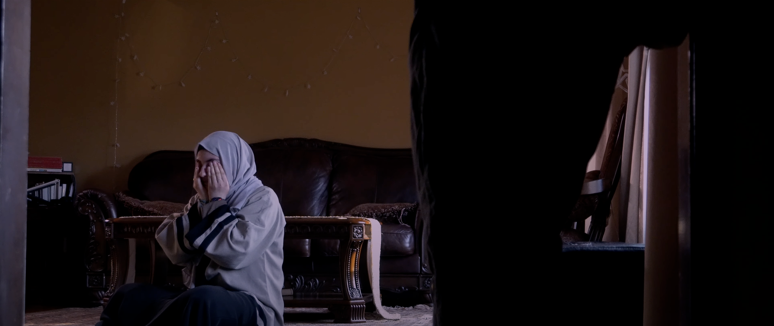 A young woman wearing a hijab sitting on the floor of a living room, covering her face with her hands, while a person stands nearby.