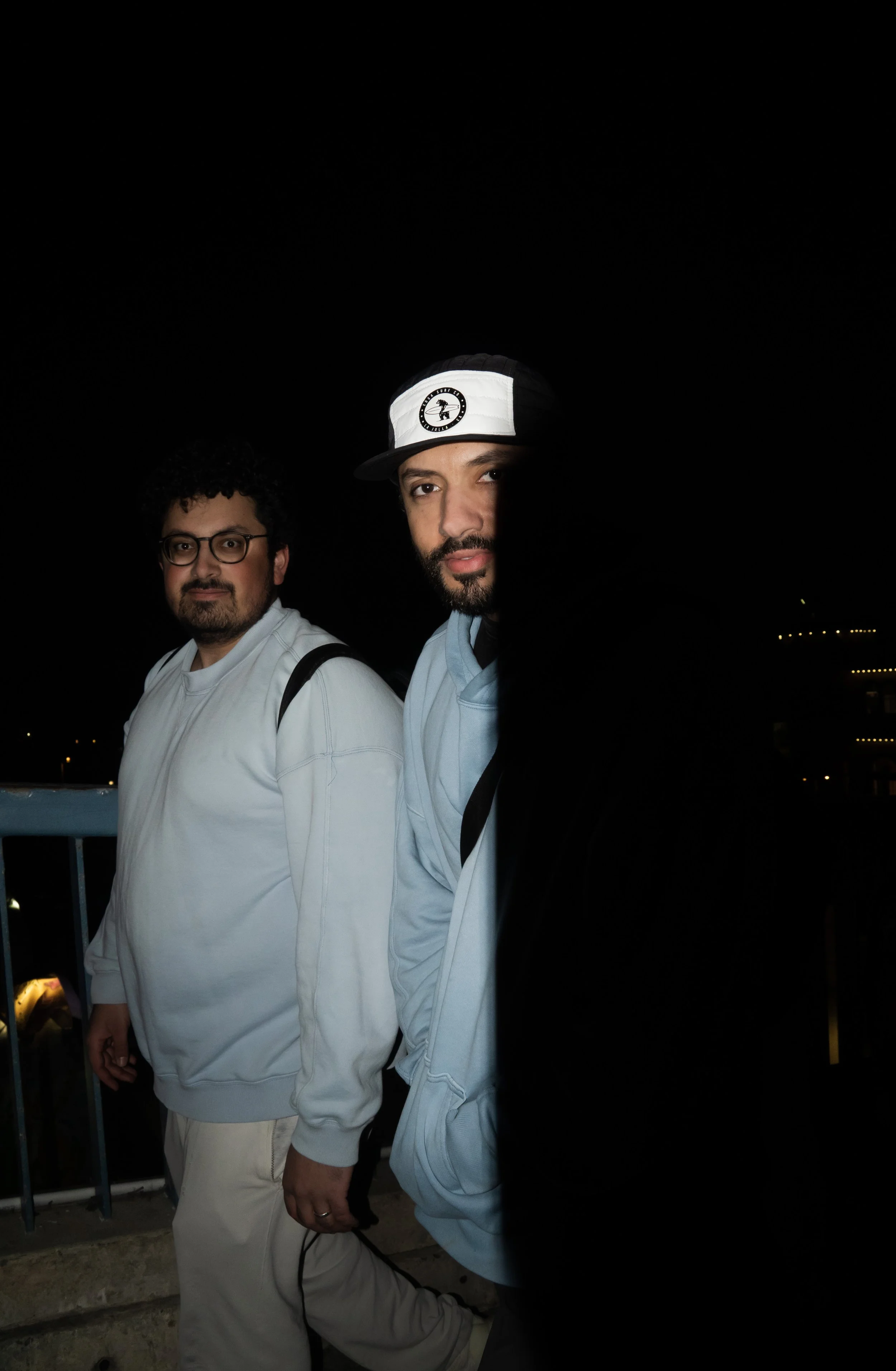 Two men standing outdoors at night, one wearing glasses and a white sweatshirt, the other wearing a light blue hoodie and a black and white hat, with a dark background and faint city lights.