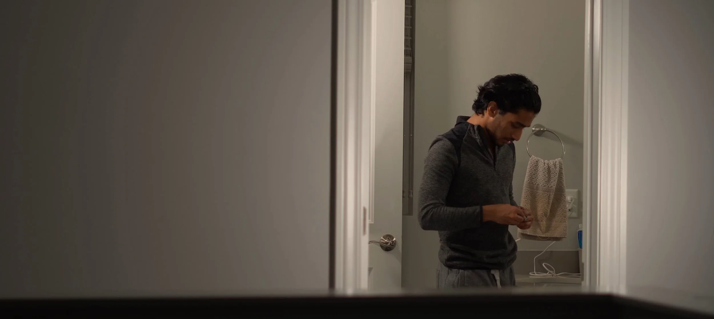 A man with dark hair is standing in a bathroom, looking down at his hands, wearing a dark gray hoodie and sweatpants.