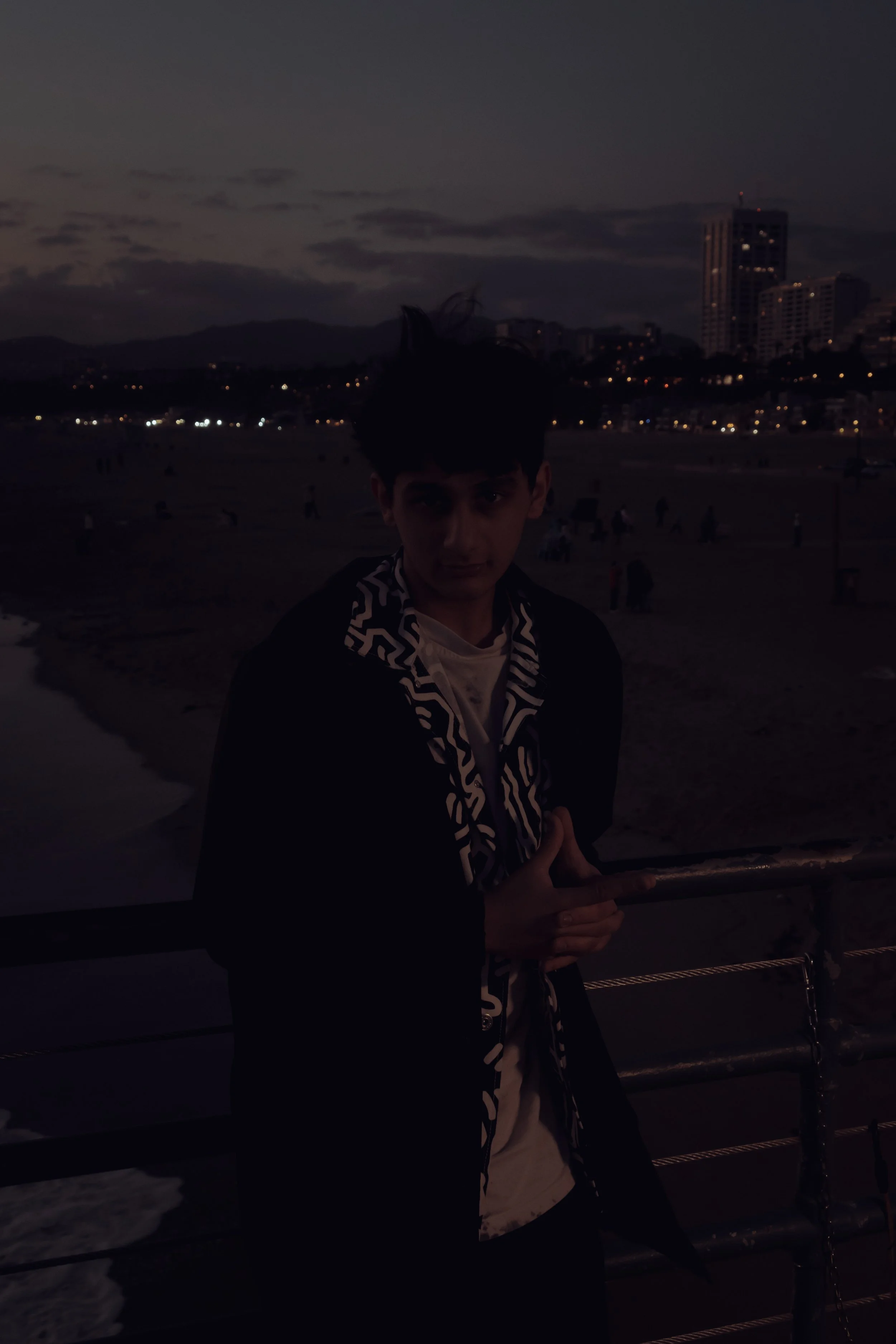A person standing near a seaside railing during dusk, with a city skyline and mountains in the background.