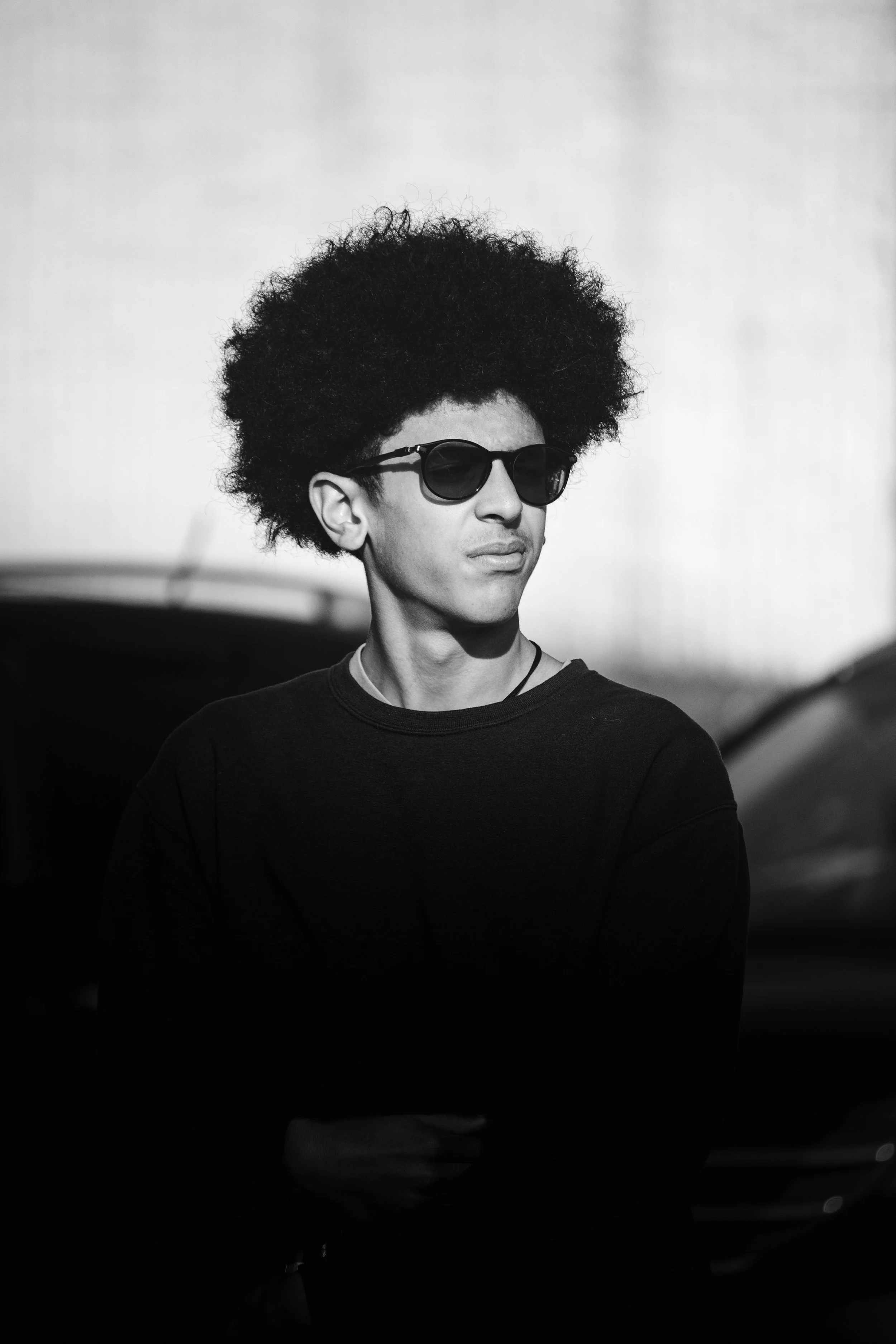Black and white photo of a young man wearing sunglasses, a black shirt, and a necklace, standing outdoors with blurred cars in the background.