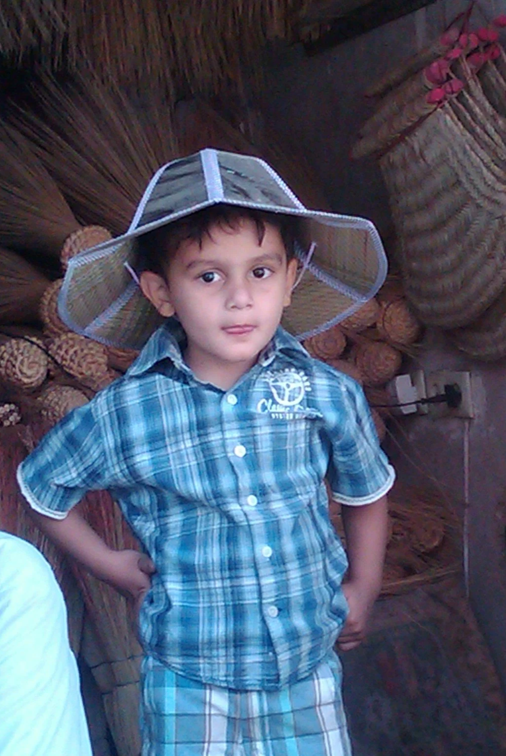 A young boy wearing a plaid blue shirt and plaid shorts, standing with hands on hips, wearing a wide-brimmed transparent hat, standing in front of a wooden background with logs and woven baskets.