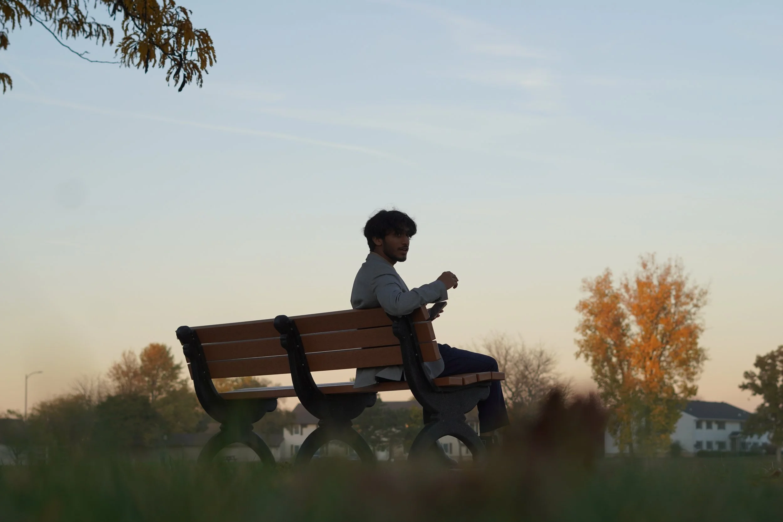 A man sitting on a park bench at sunset, holding a phone and looking over his shoulder.