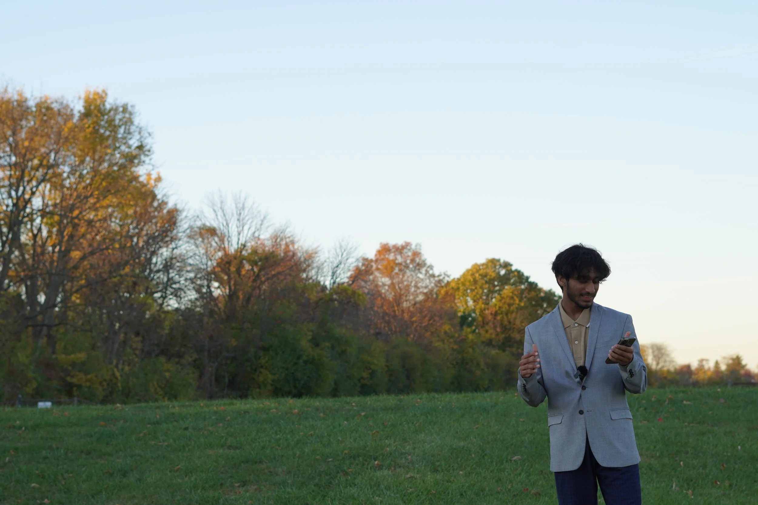 Man in gray blazer standing on grassy field using smartphone with autumn trees in background.
