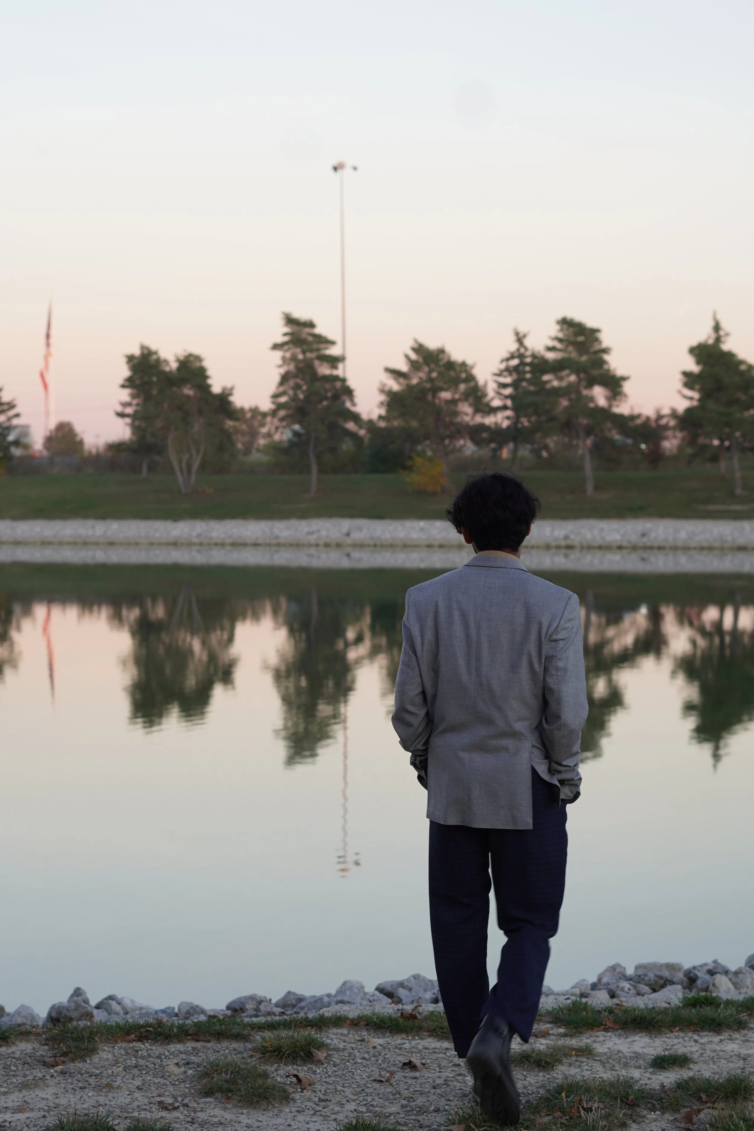A person in a gray blazer and dark pants walks near a lake during sunset, reflecting trees and a flag in the water.