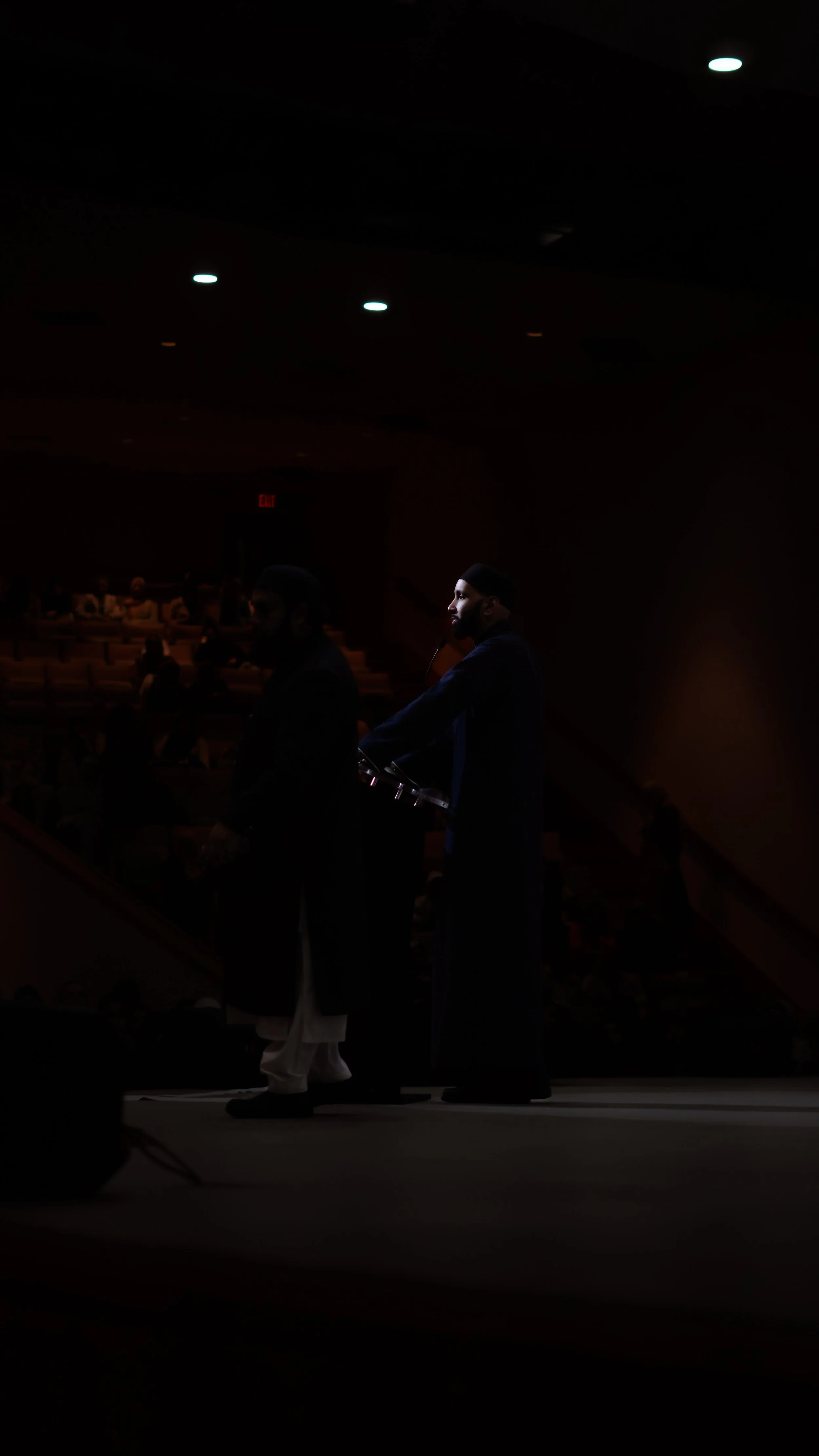 Two men in traditional attire standing on stage, with a dark audience in the background, under dim lighting.