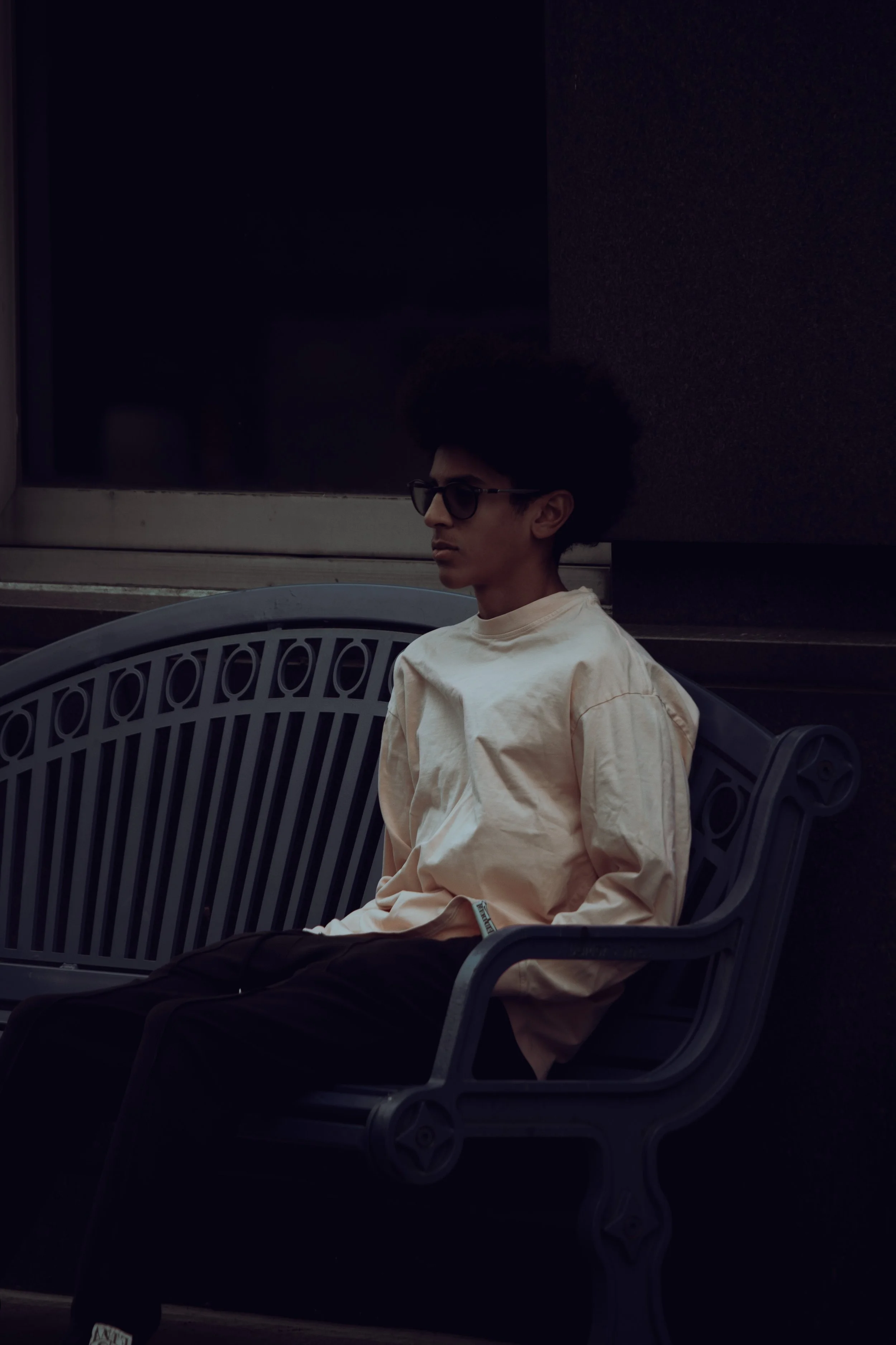 A young man with dark, curly hair wearing sunglasses, a beige oversized sweatshirt, and black pants is sitting alone on a black park bench in front of a dark building, looking to the side with a serious expression.