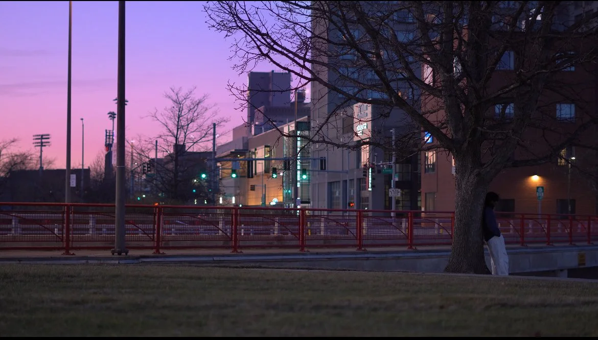 A person leaning against a tree on a sidewalk in an urban area during dusk with a purple sky, buildings, streetlights, and traffic signals visible in the background.