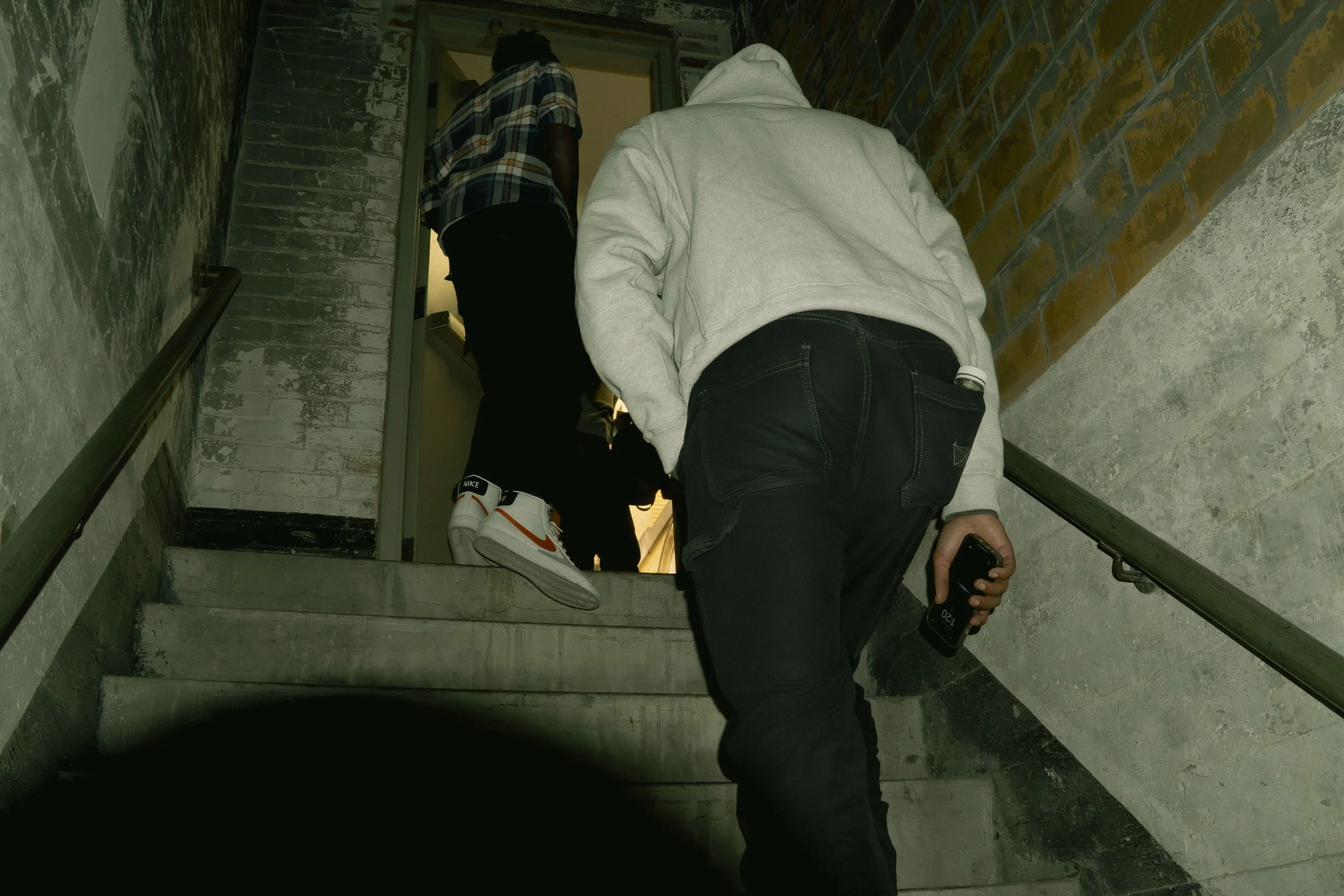 Two people ascending a worn, industrial-style staircase with concrete steps and brick walls. One individual is wearing a white hoodie and black pants, holding a phone. The other is wearing a checkered shirt and sneakers, walking ahead.