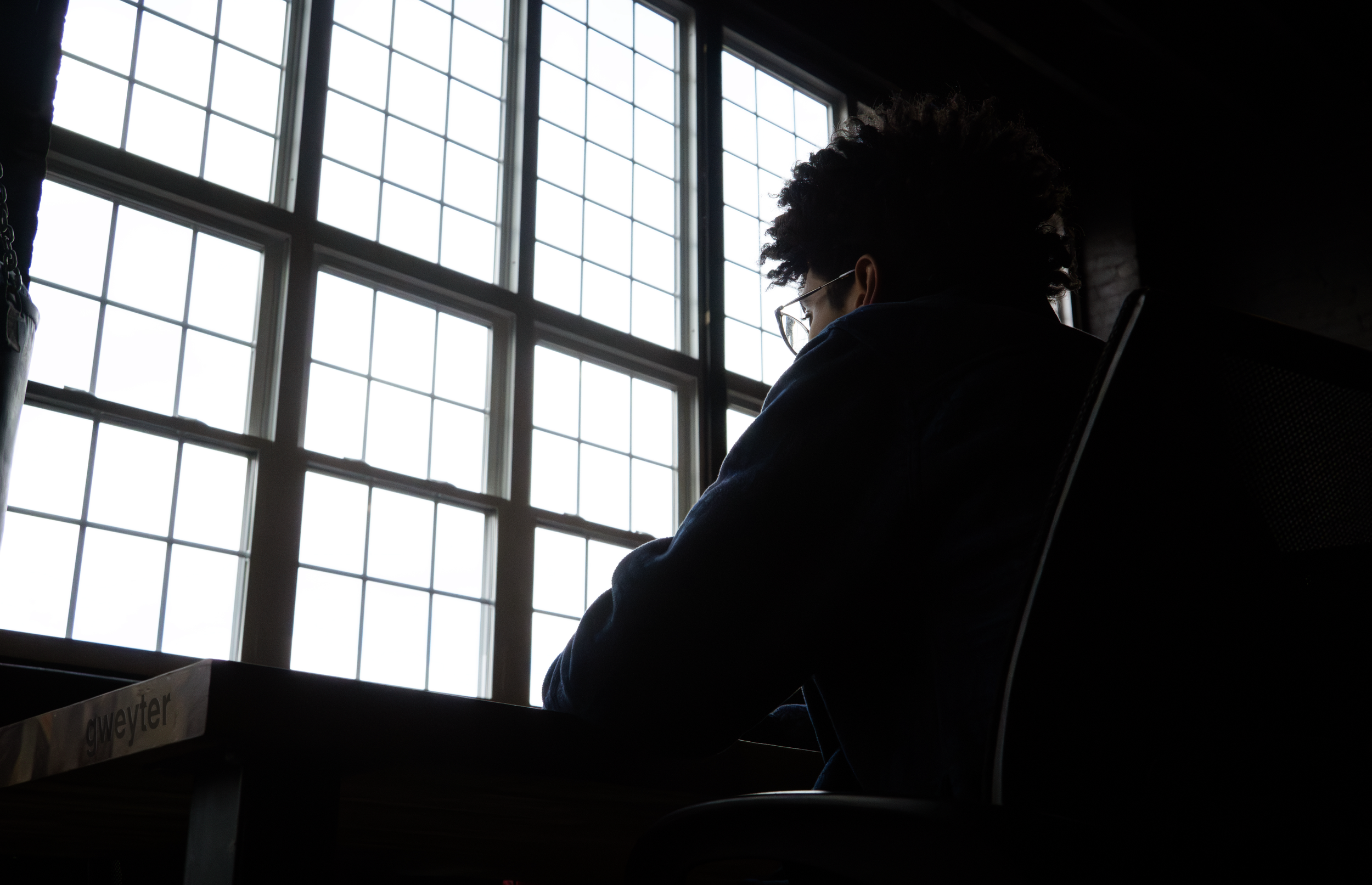 A person sitting in a dark room near a large window, looking outside. The person has glasses and curly hair, with their silhouette visible against the bright window.