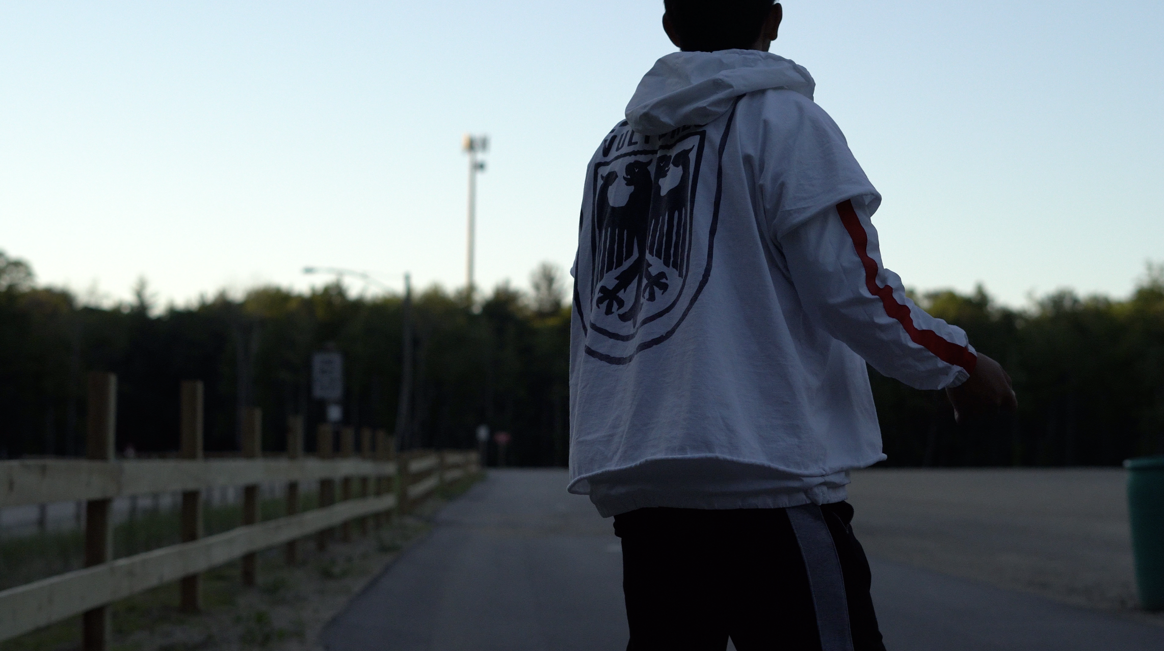 A person walking outdoors on a trail with a fence, wearing a white jacket with a graphic on the back, in the early evening or late afternoon with the sky fading.