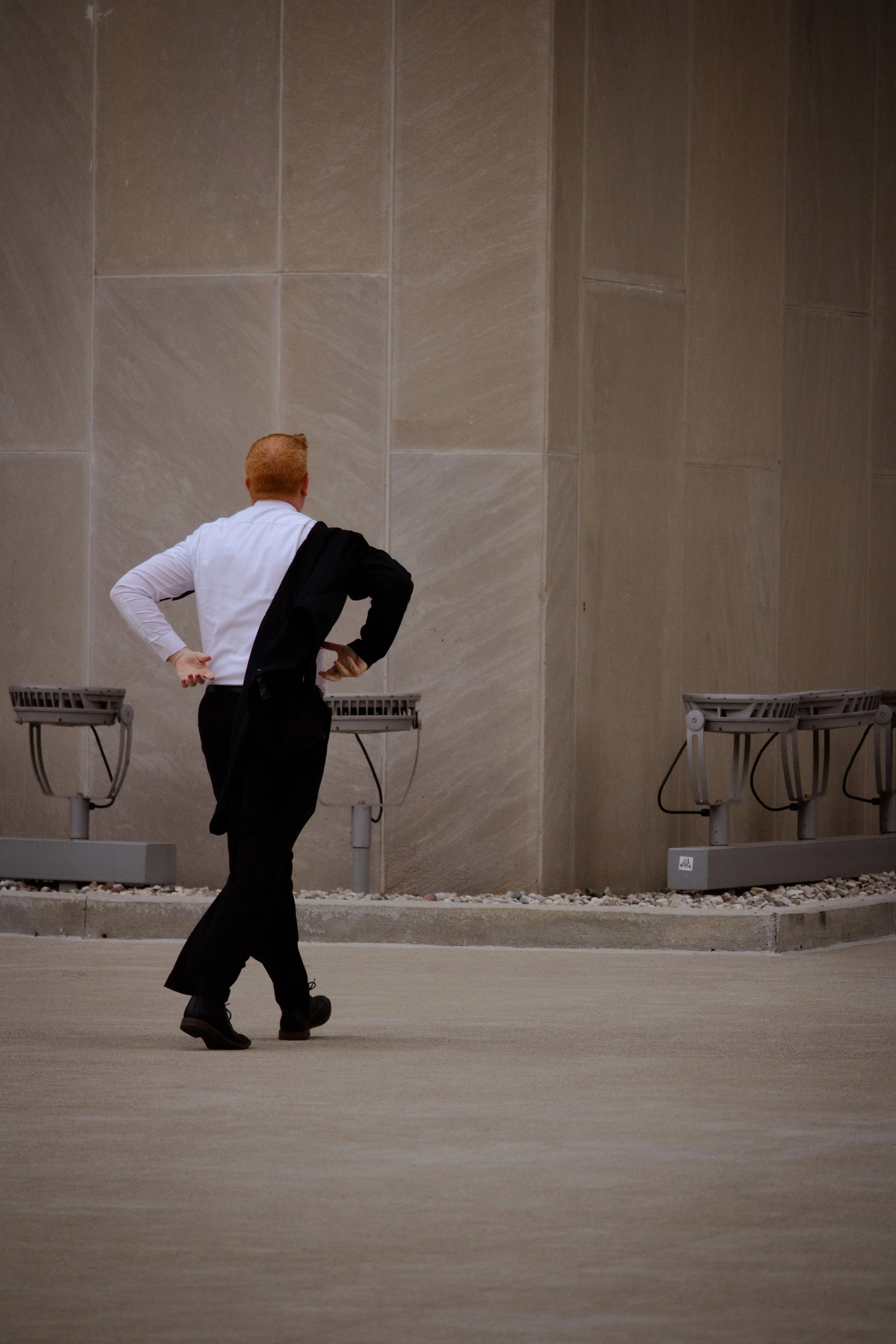 A person with red hair, wearing a white shirt and black pants, is walking away from the camera with a black jacket draped over their shoulder, in an indoor space with beige tiled walls and metal benches.