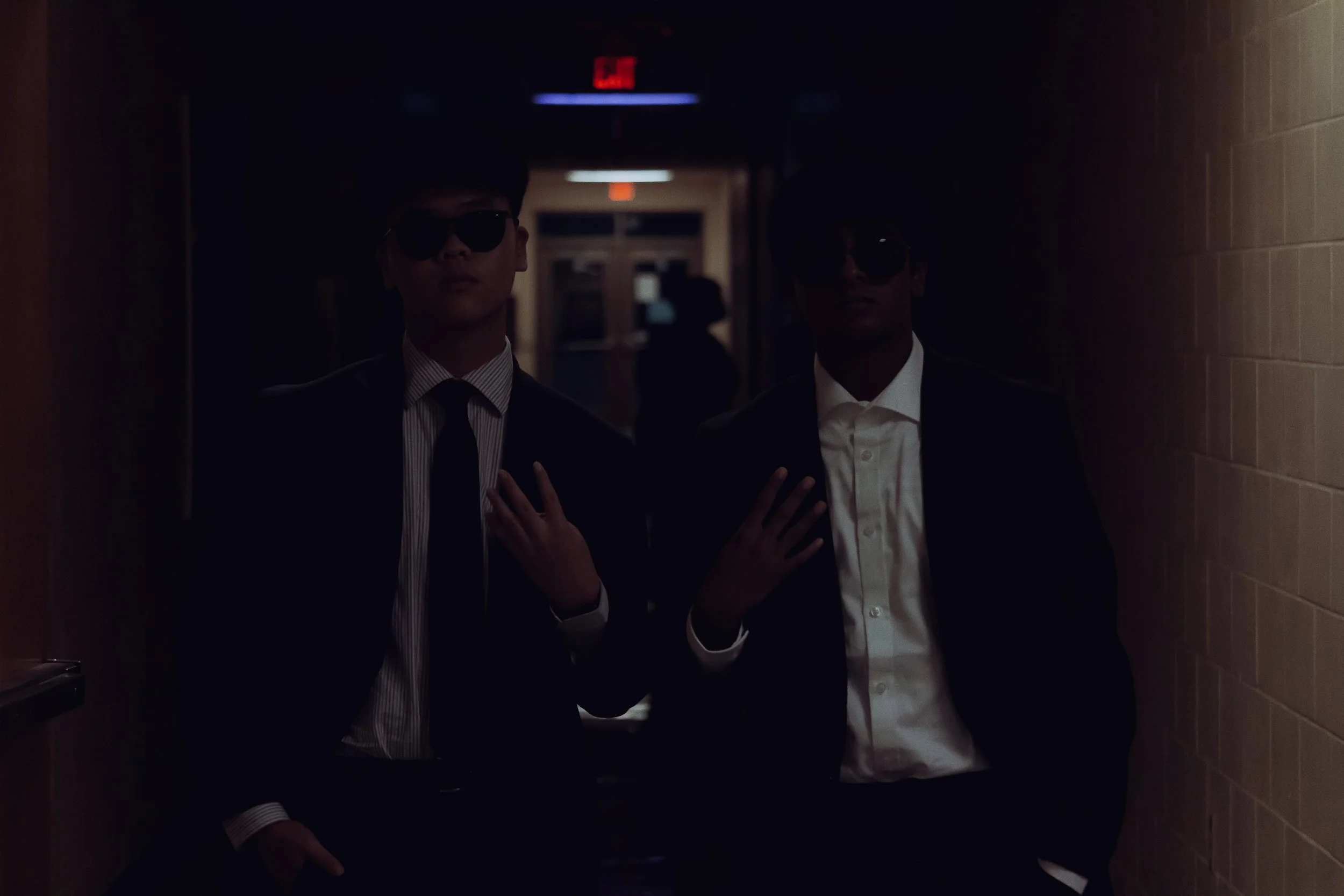 Two young men in suits and sunglasses standing in a dark hallway with a faintly lit background.