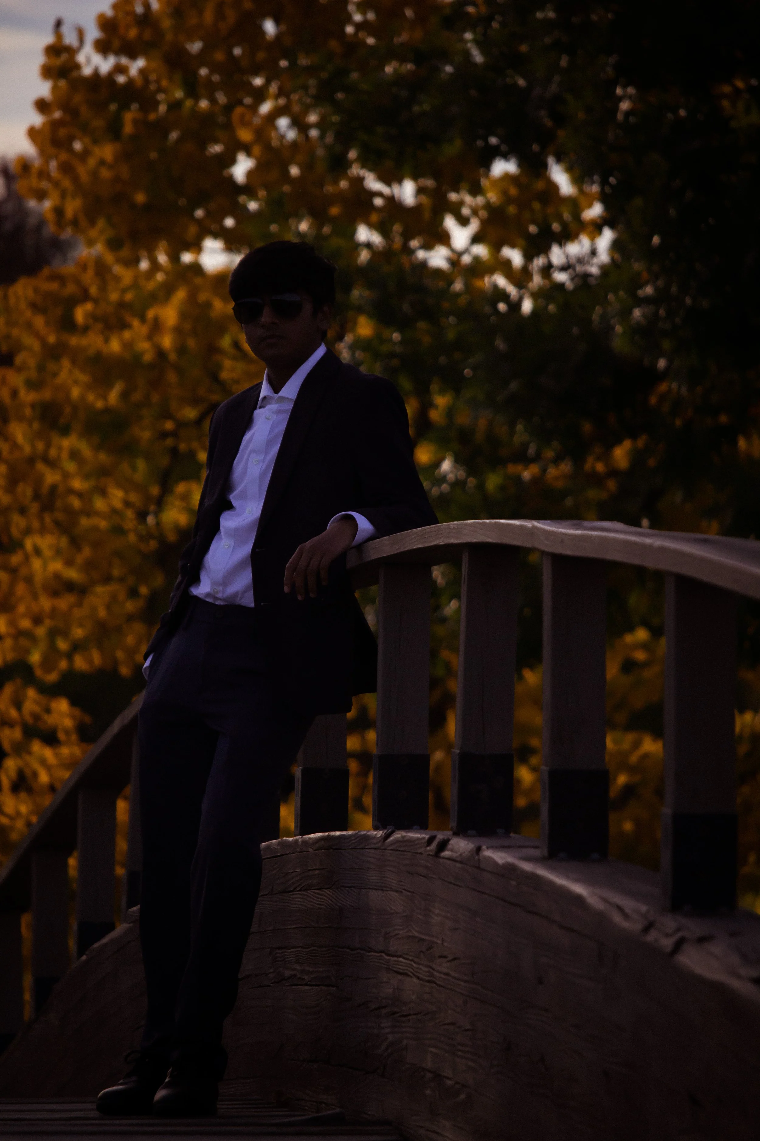 A man in a black suit, white shirt, and sunglasses leaning on a wooden bridge railing during sunset, with orange and yellow autumn trees in the background.