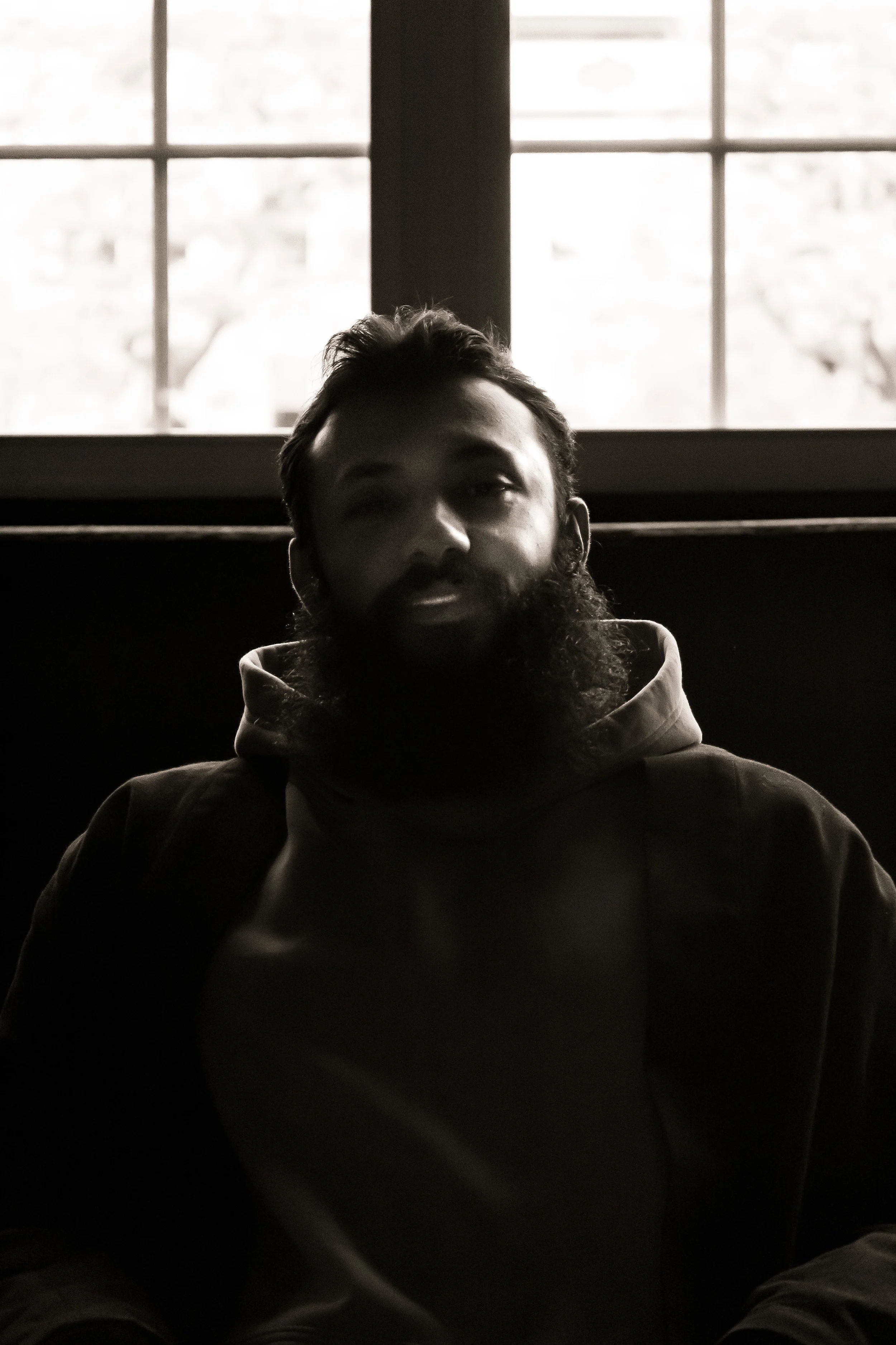 A man with a beard sitting indoors near a window, appears to be in a dimly lit room with the light coming from behind.