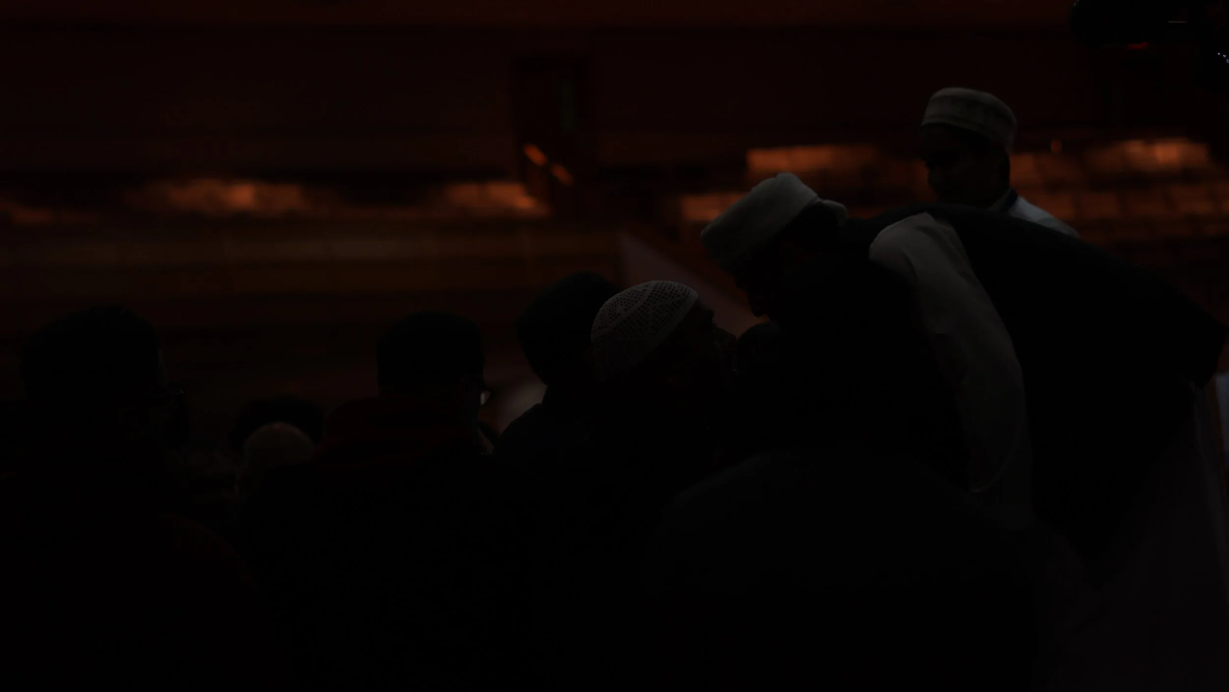 Silhouettes of men in traditional clothing, some wearing white caps, in a dark setting with a dimly lit, wooden interior background.