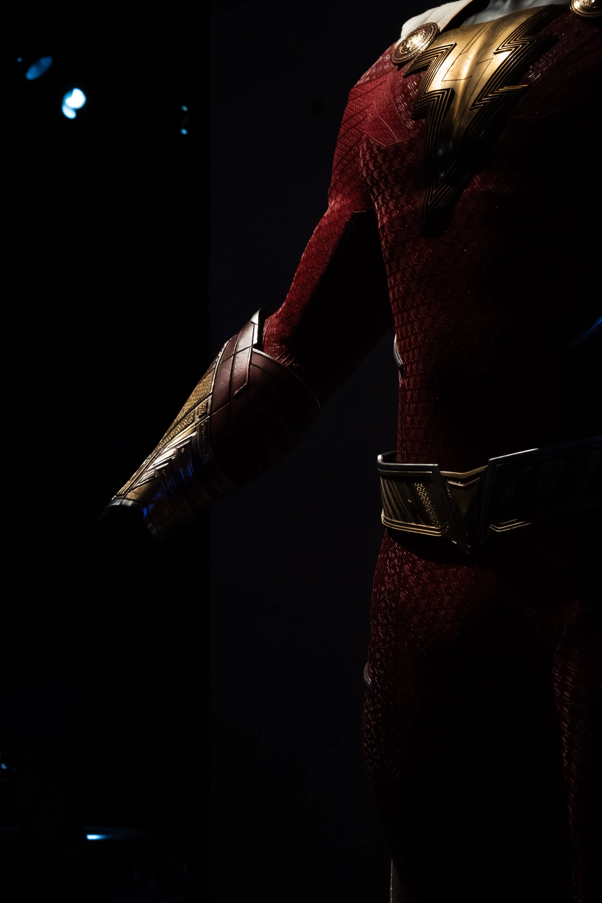 Close-up of Wonder Woman's costume, showing her red textured bodysuit, gold armor accents on her shoulder and waist, and her left arm extended with a gold and black bracer.