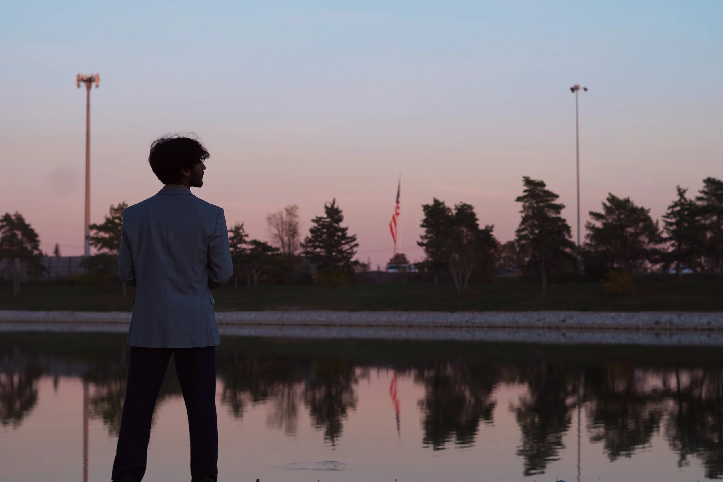 A man in a gray blazer standing by a body of water during sunset, with trees, flagpoles, and an American flag in the background.