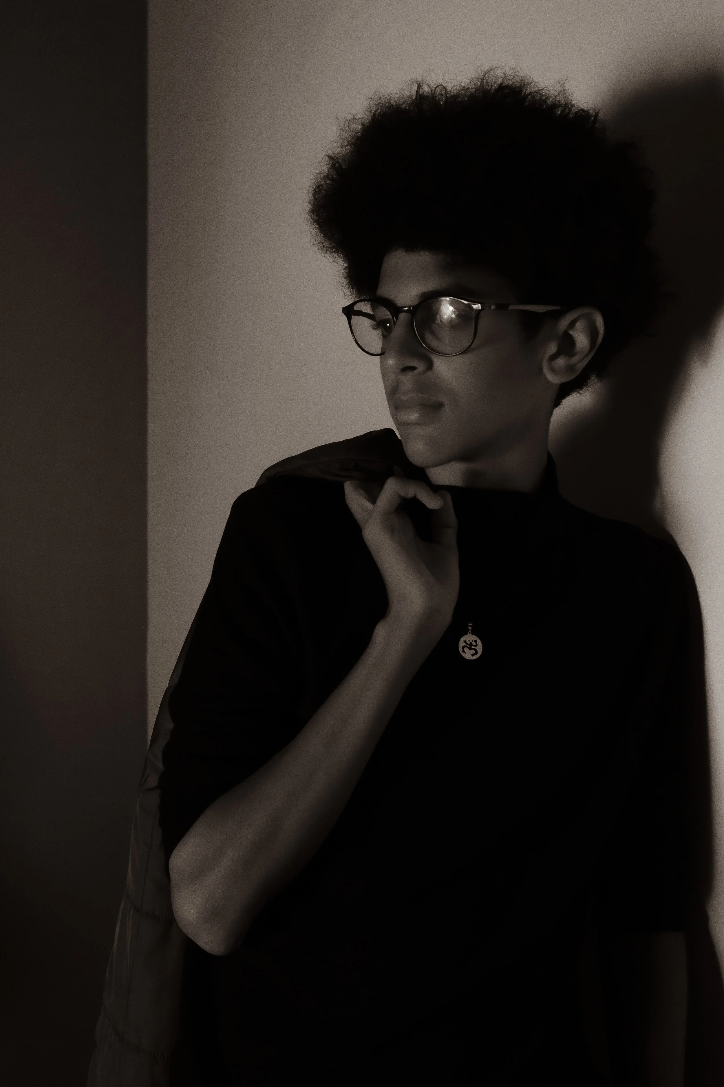 A young person with curly hair and glasses, holding an jacket over their shoulder, looking thoughtfully to the side, in a dark, moody setting with shadows.